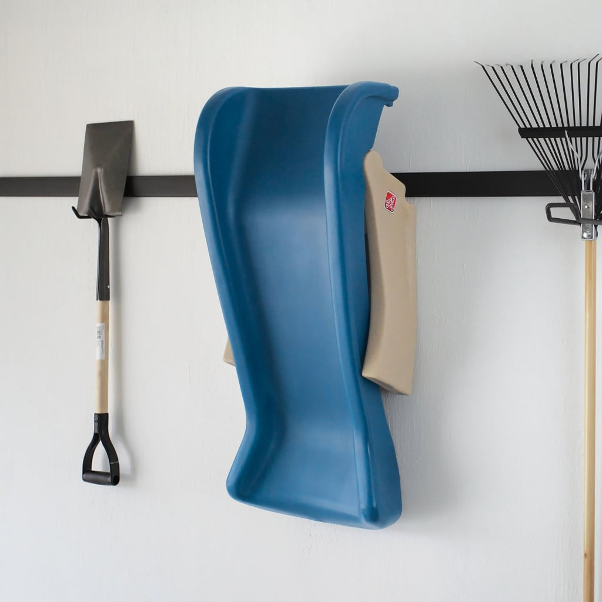 Eine blaue Rutsche hängt an einer weißen Wand mit einer schwarzen Stange. Eine Schaufel und eine Harke hängen ebenfalls an der Wand.
