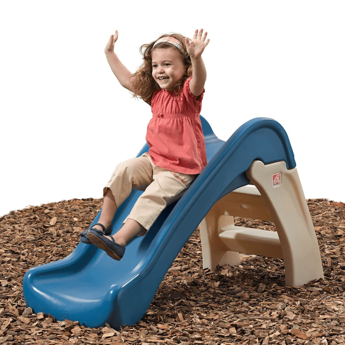 Ein junges Mädchen rutscht eine blaue Plastikrutsche hinunter und hebt die Arme. Sie trägt ein rotes Oberteil, beige Hose und ein Stirnband. Die Rutsche steht auf Holzschnitzeln.