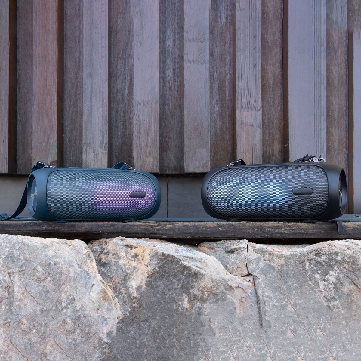 Two portable speakers on a rock. They are black and teal, with a wooden background. The speakers have carrying straps.
