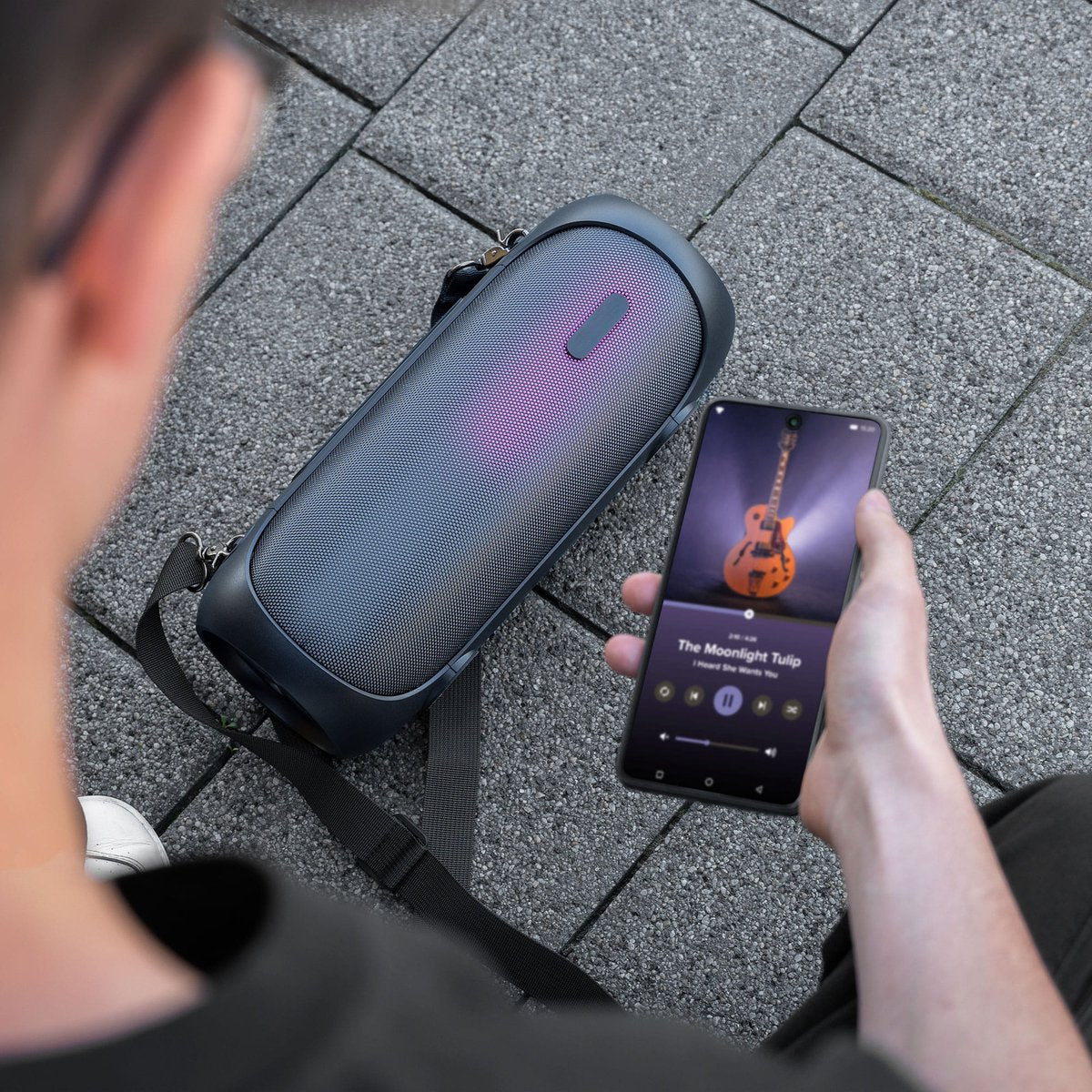 Person holding a phone playing music, next to a dark speaker. The speaker has a strap and is sitting on a stone surface.