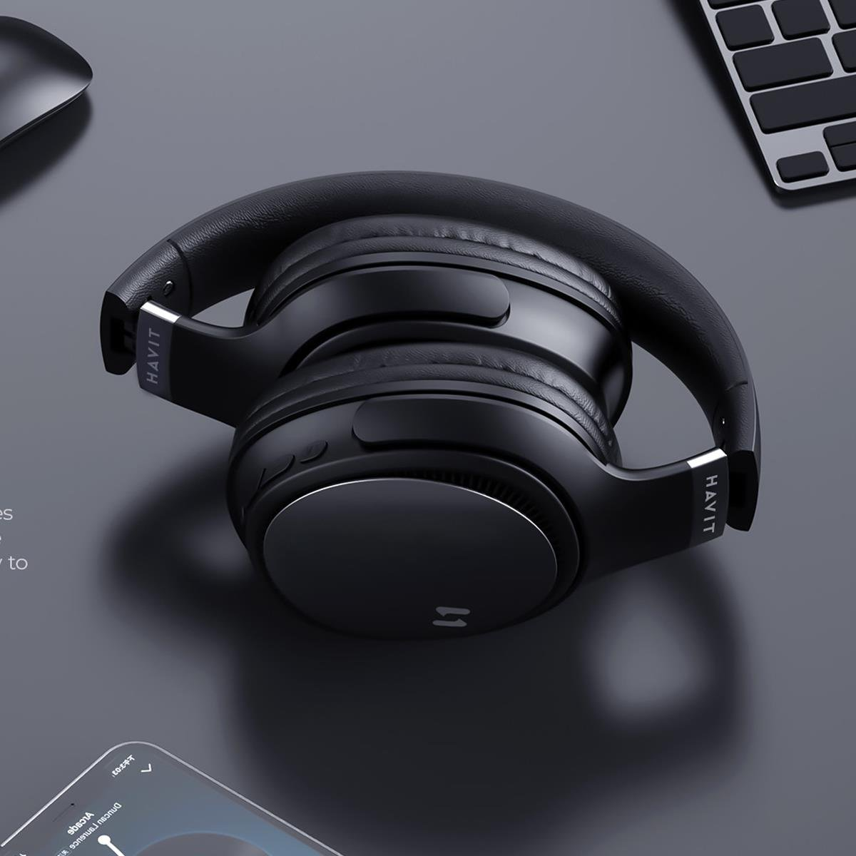 Auriculares HAVIT negros doblados sobre una superficie gris, con un ratón y un teclado en el fondo. Los auriculares tienen un acento plateado.