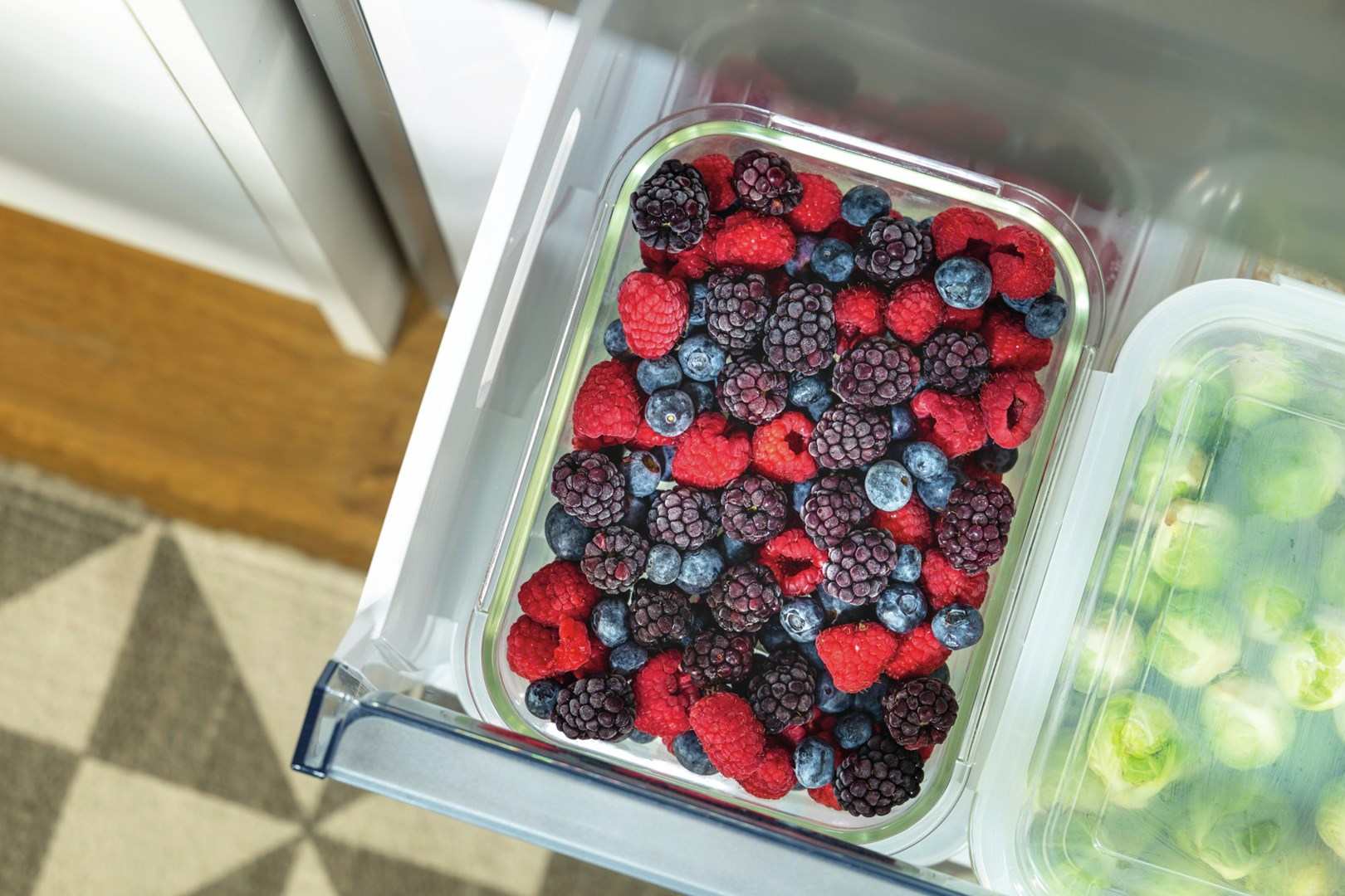 Szuflada lodówki wypełniona mieszanymi jagodami: malinami, jeżynami i borówkami. Drugi pojemnik zawiera brukselkę.