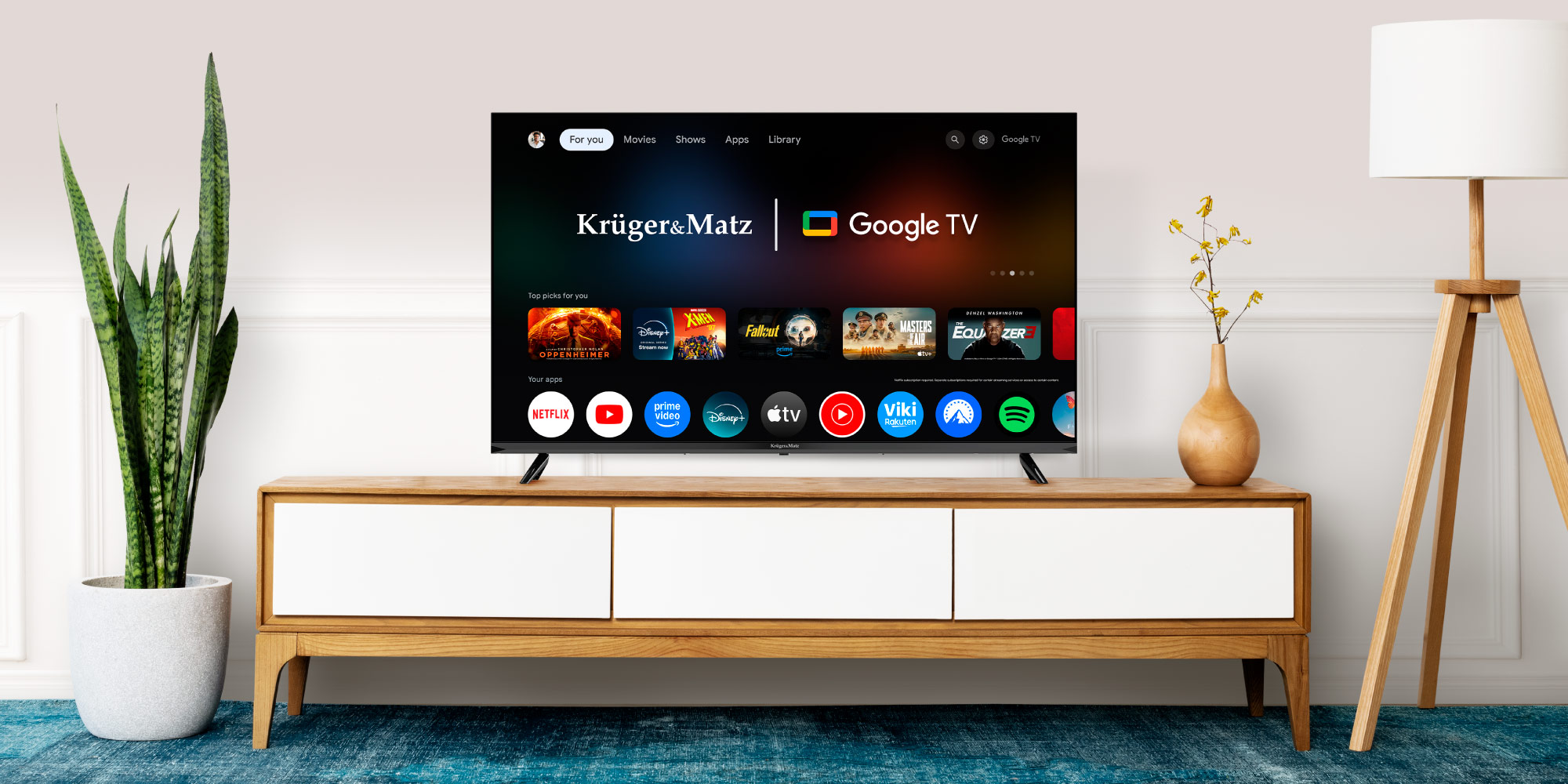 Telewizor wyświetla interfejs Google TV. Jest na drewnianej szafce z rośliną i lampą w pobliżu. Poniżej znajduje się niebieski dywan.