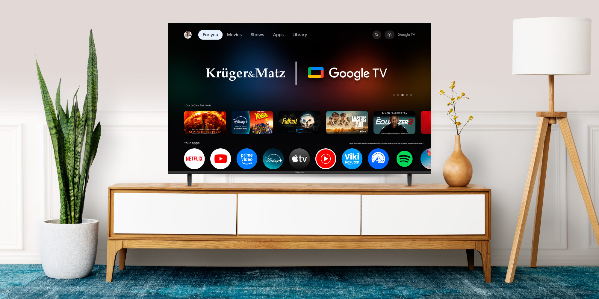 Telewizor wyświetla interfejs Google TV. Stoi na drewnianej szafce, z rośliną i lampą w pobliżu. Niebieski dywan leży na podłodze.