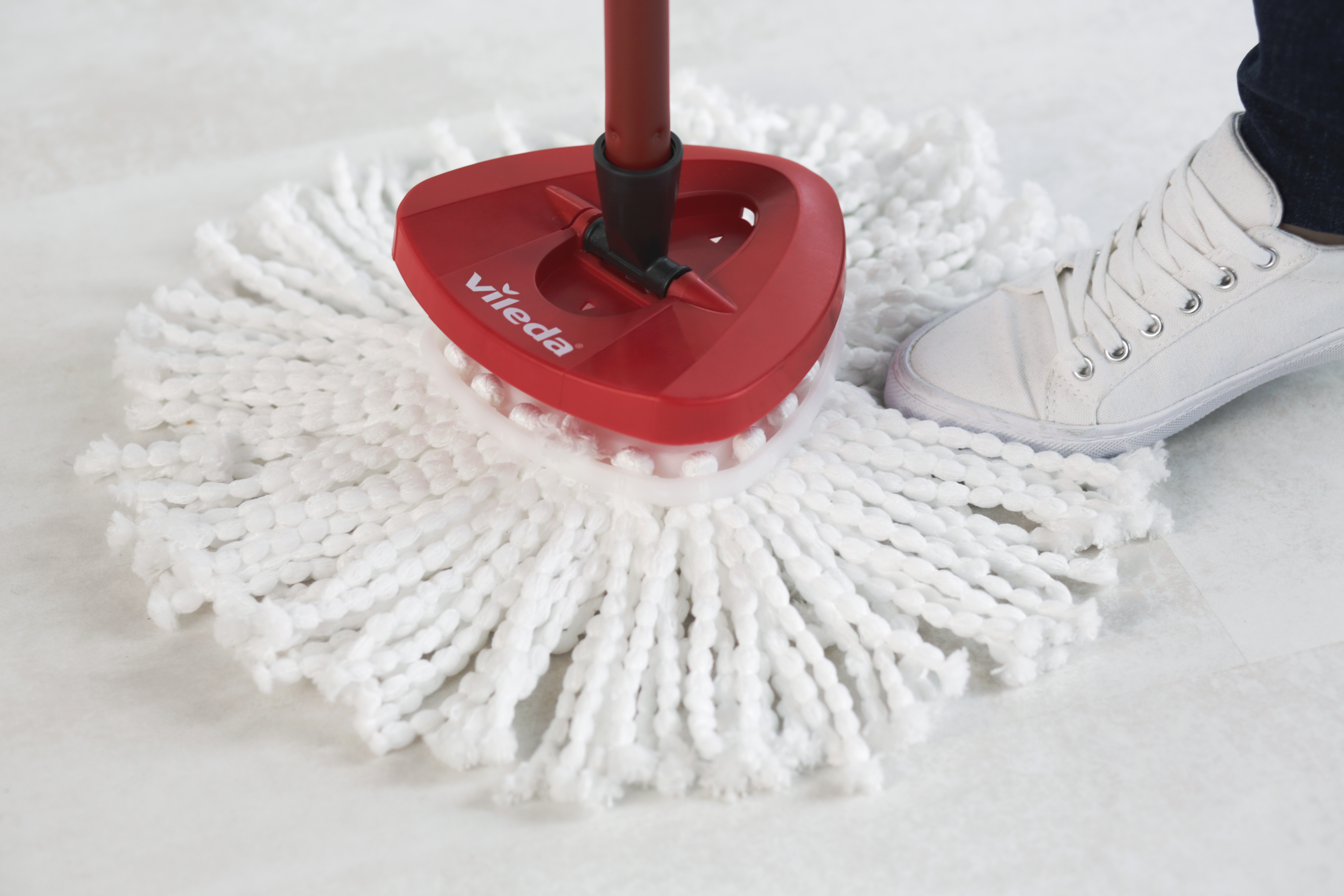 Una mopa Vileda roja en un suelo blanco, con un zapato blanco visible. La mopa tiene un cabezal blanco y un mango rojo.