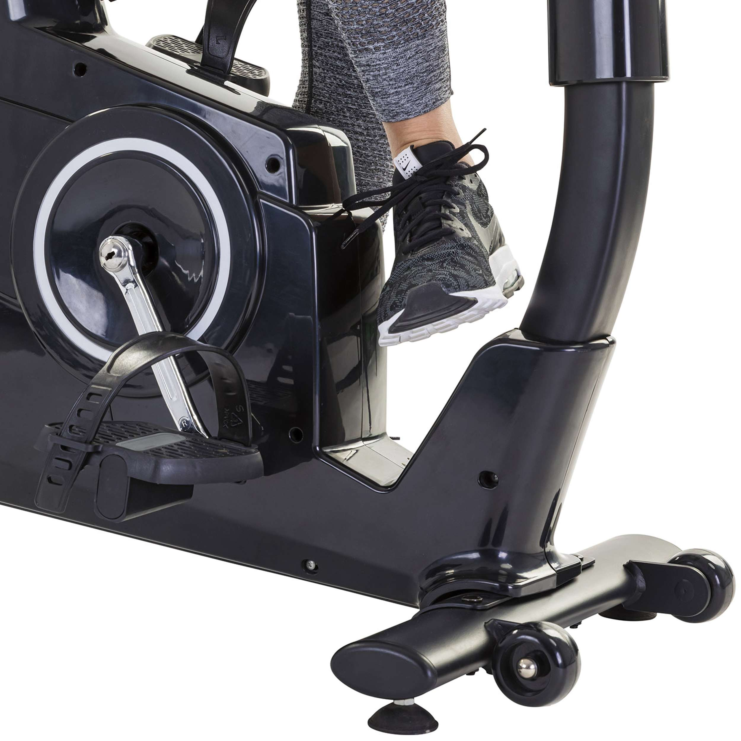 Voet van een persoon in een zwarte sportschoen op het pedaal van een zwarte hometrainer. Een grijze trainingsbroek is ook zichtbaar.