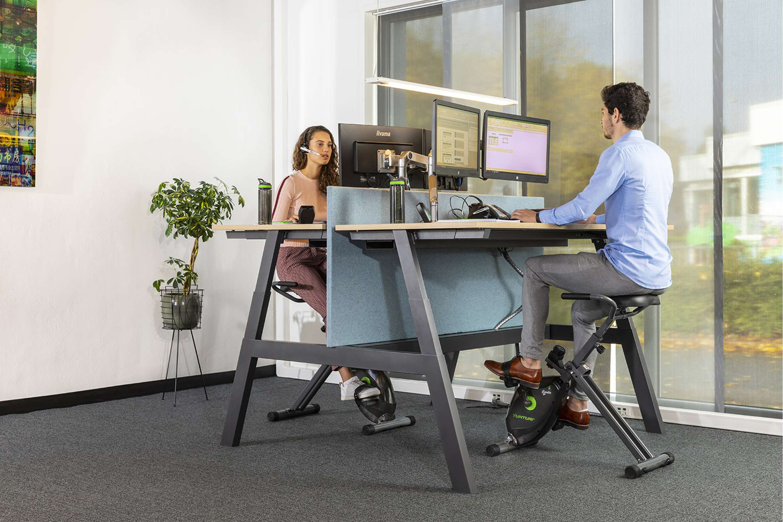 Twee mensen werken aan bureaus met hometrainers. Eén persoon draagt een headset. Monitors op bureaus. Planten en raam.