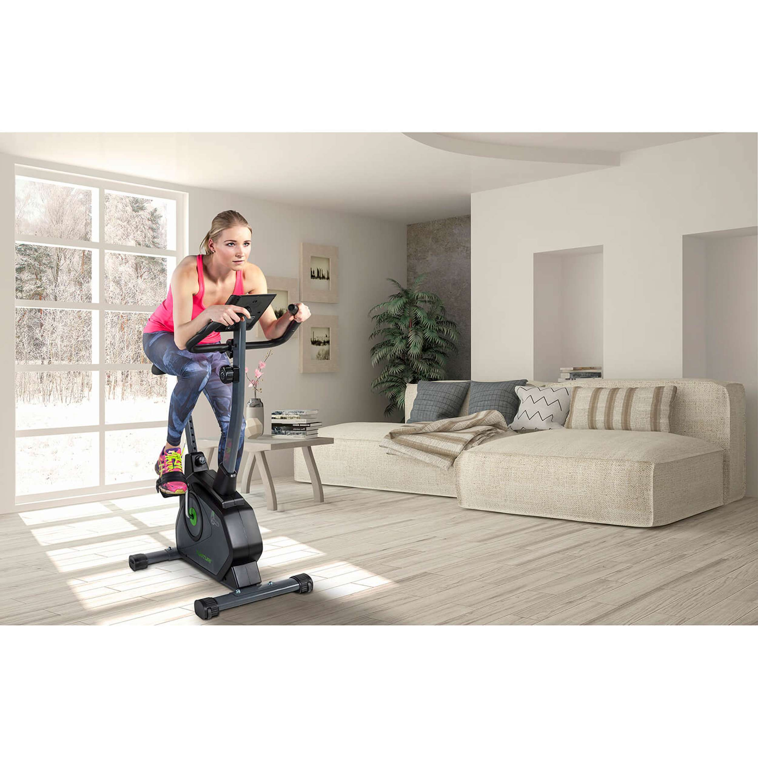 Woman on exercise bike in a bright room. Background: window with snowy view, beige couch, and wooden floor.