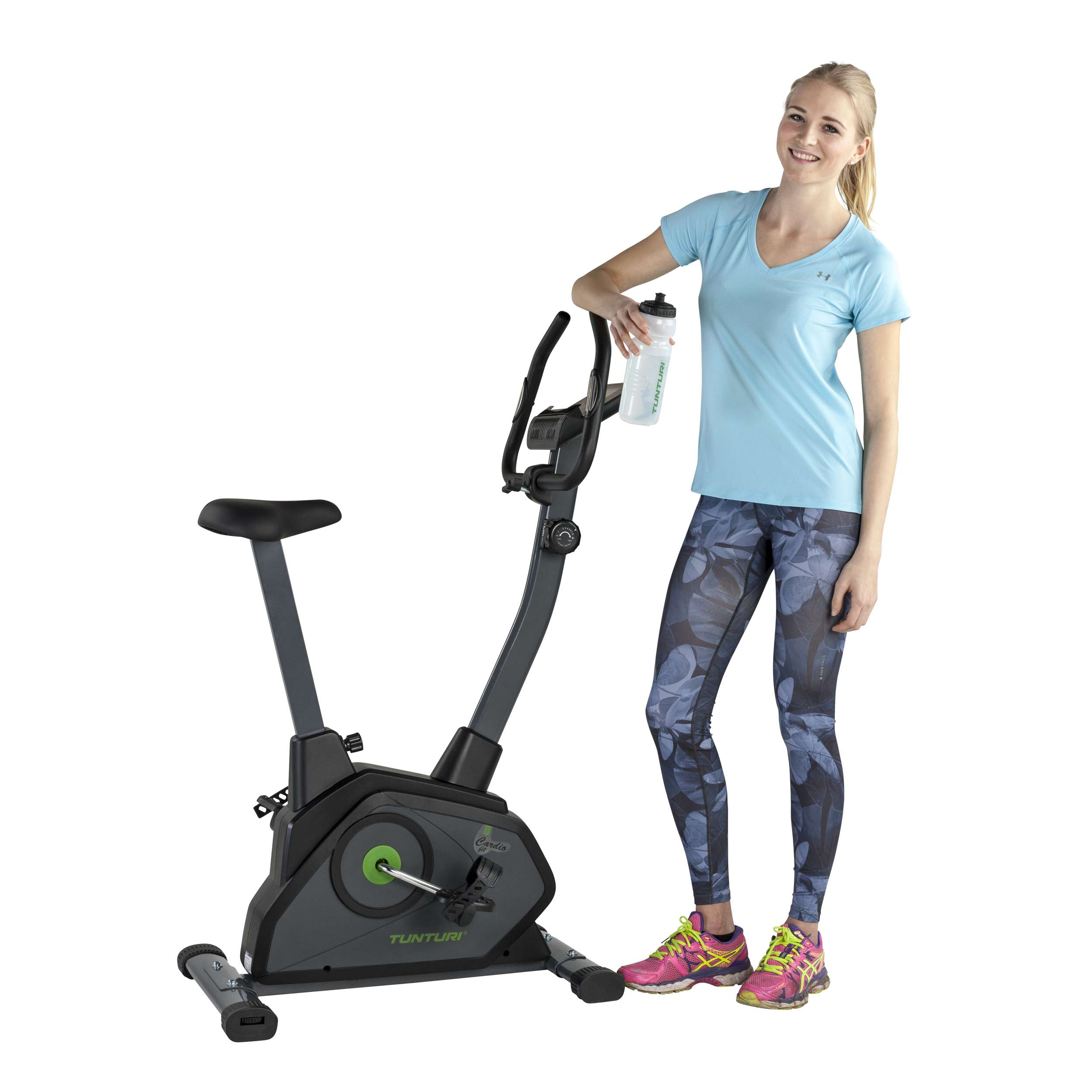 A woman in athletic wear stands near an exercise bike, holding a water bottle. She smiles, with the bike on the left.