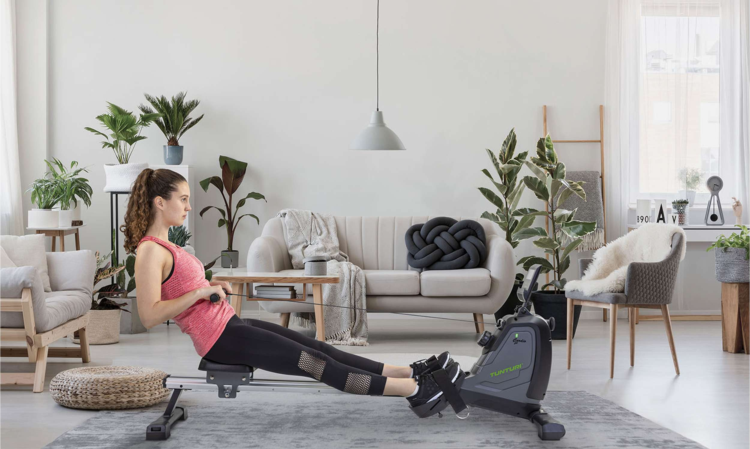 Een vrouw traint op een roeimachine in een woonkamer. Planten, een bank en een raam met gordijnen staan op de achtergrond.