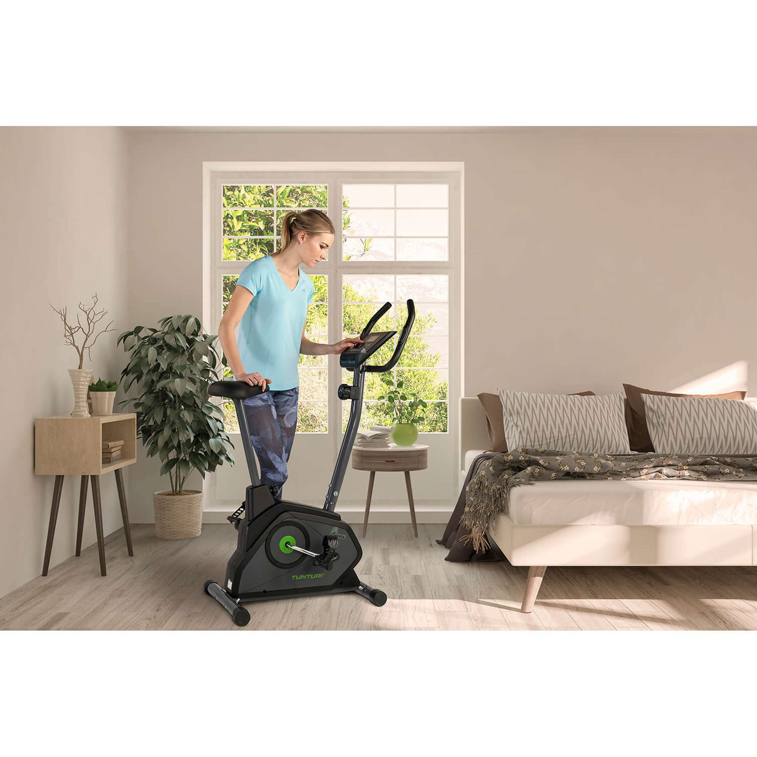 Een vrouw in een blauw shirt gebruikt een hometrainer. Een bed en een raam staan op de achtergrond. Planten en een bijzettafel zijn zichtbaar.