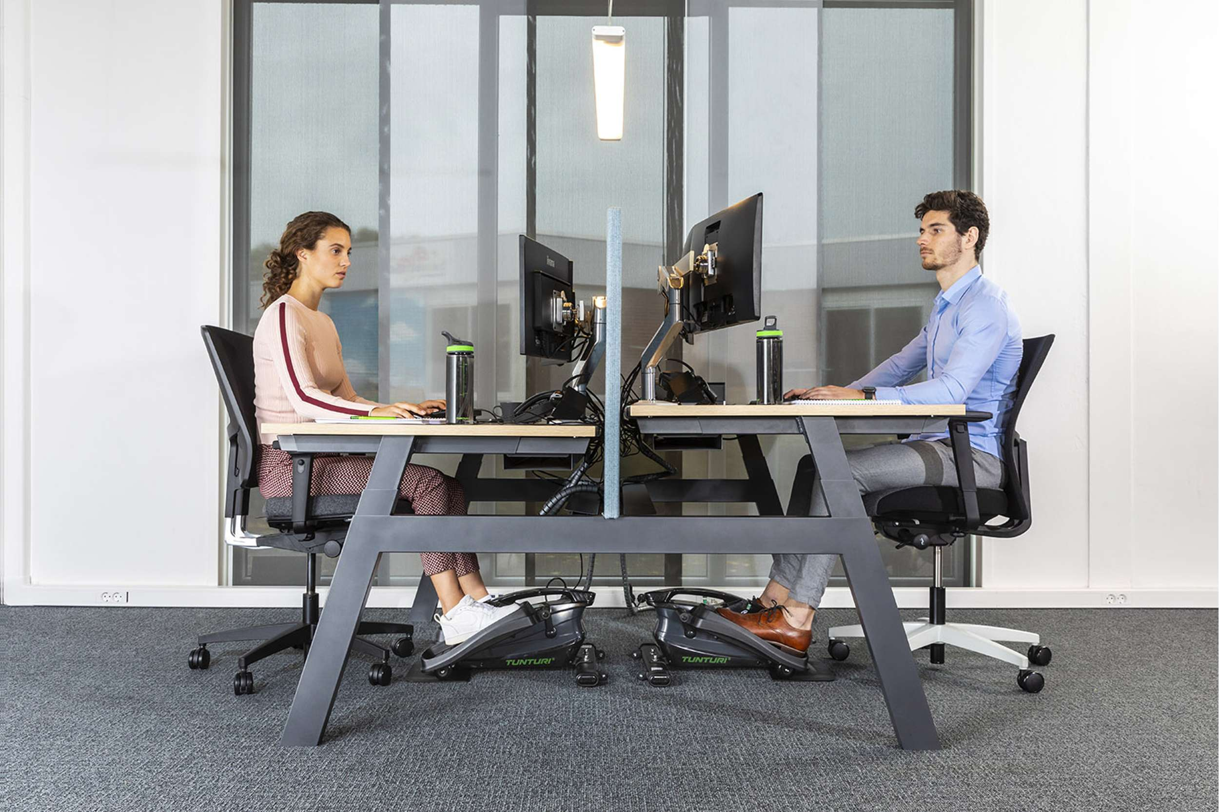 Twee mensen werken aan bureaus met computers, waterflessen en voetpedalen. De een draagt roze, de ander een blauw shirt.