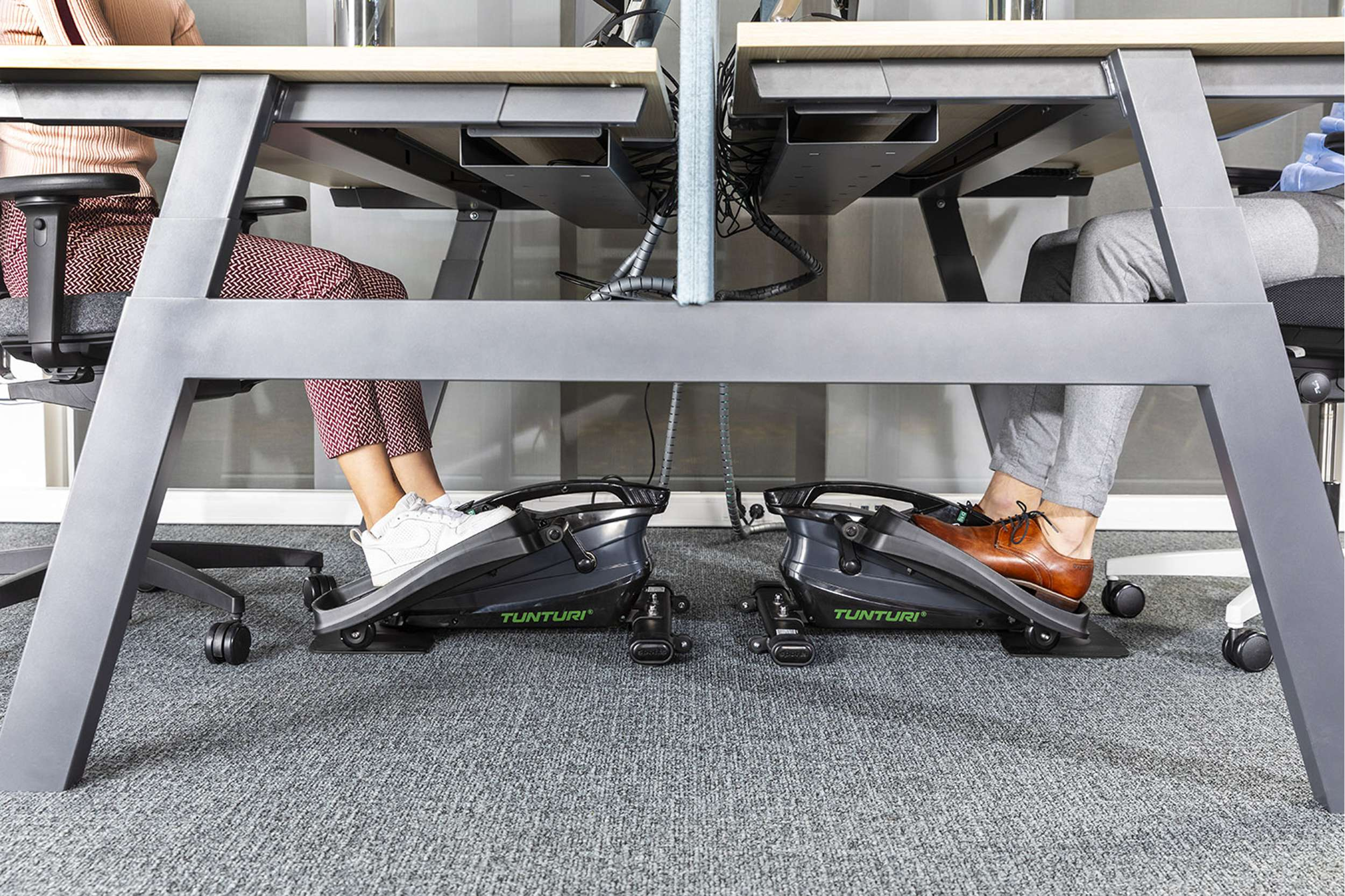 Mensen gebruiken onder-bureau trainingsmachines. Benen en voeten zijn zichtbaar, met sneakers en geklede schoenen. Bureaus, stoelen en tapijt.