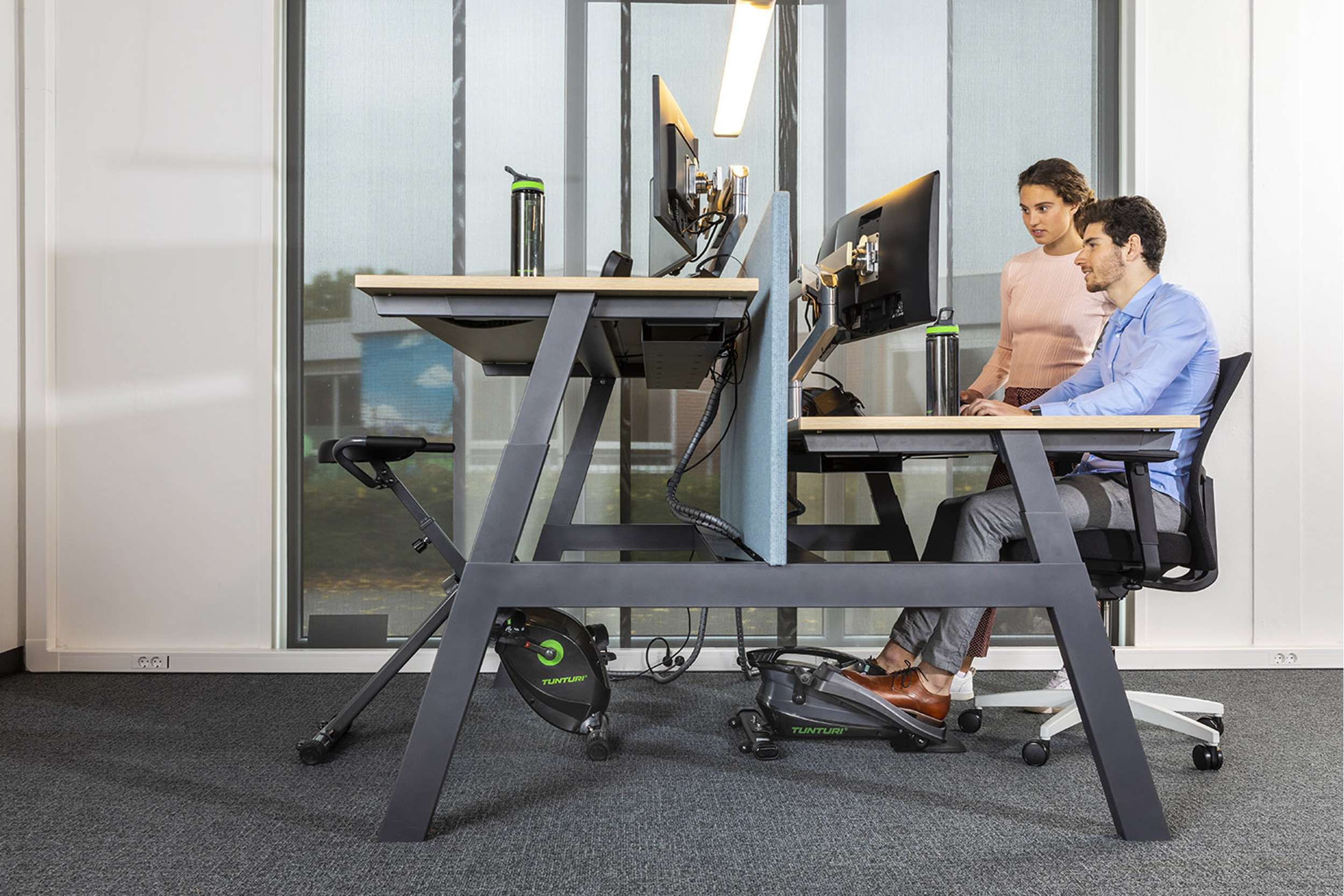 Kantoor met twee personen aan een bureau met fitnessapparatuur. Monitoren, waterflessen en een raam zijn zichtbaar.