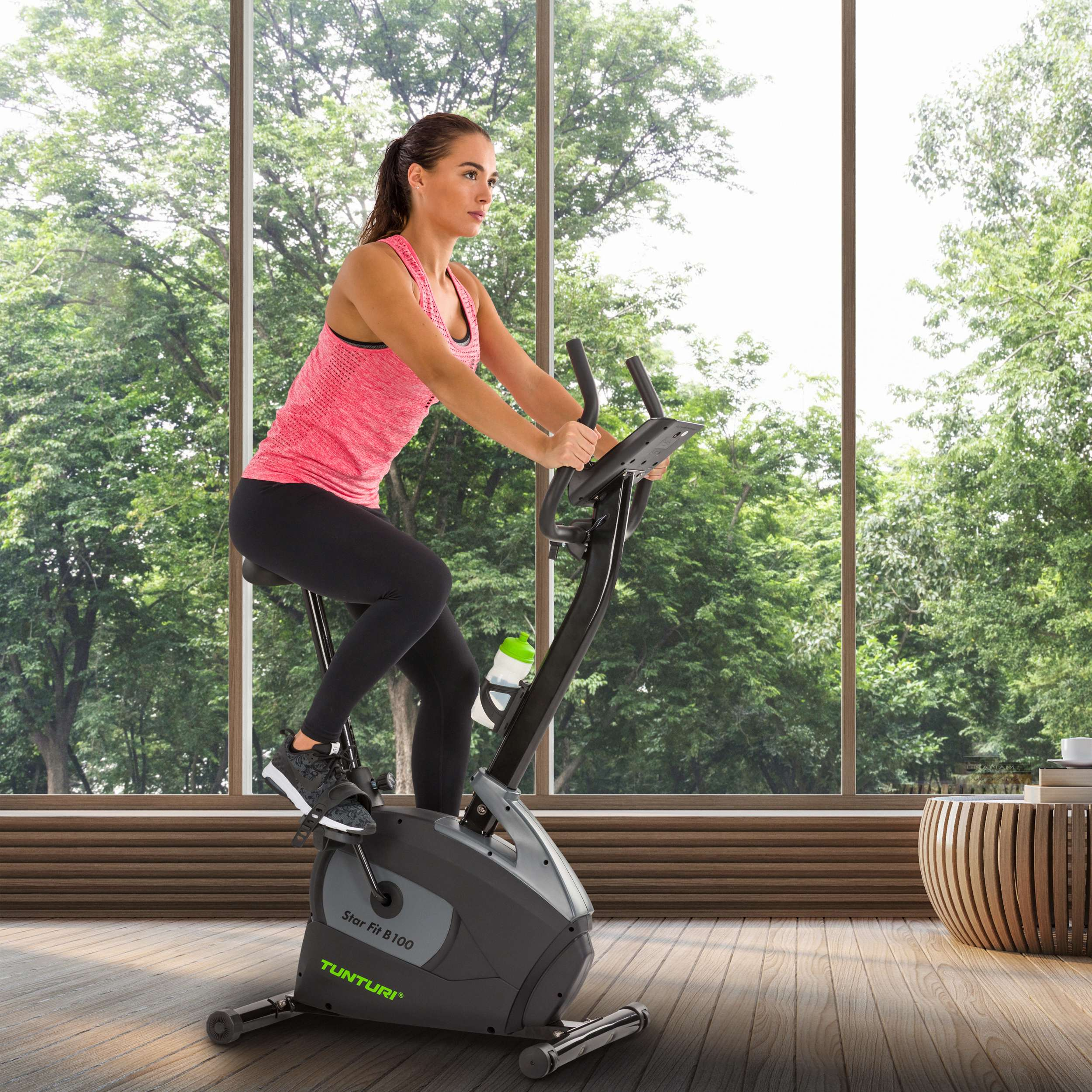 Een vrouw op een hometrainer in een kamer met een groot raam met uitzicht op bomen. De fiets is een Tunturi Star Fit B100 model.