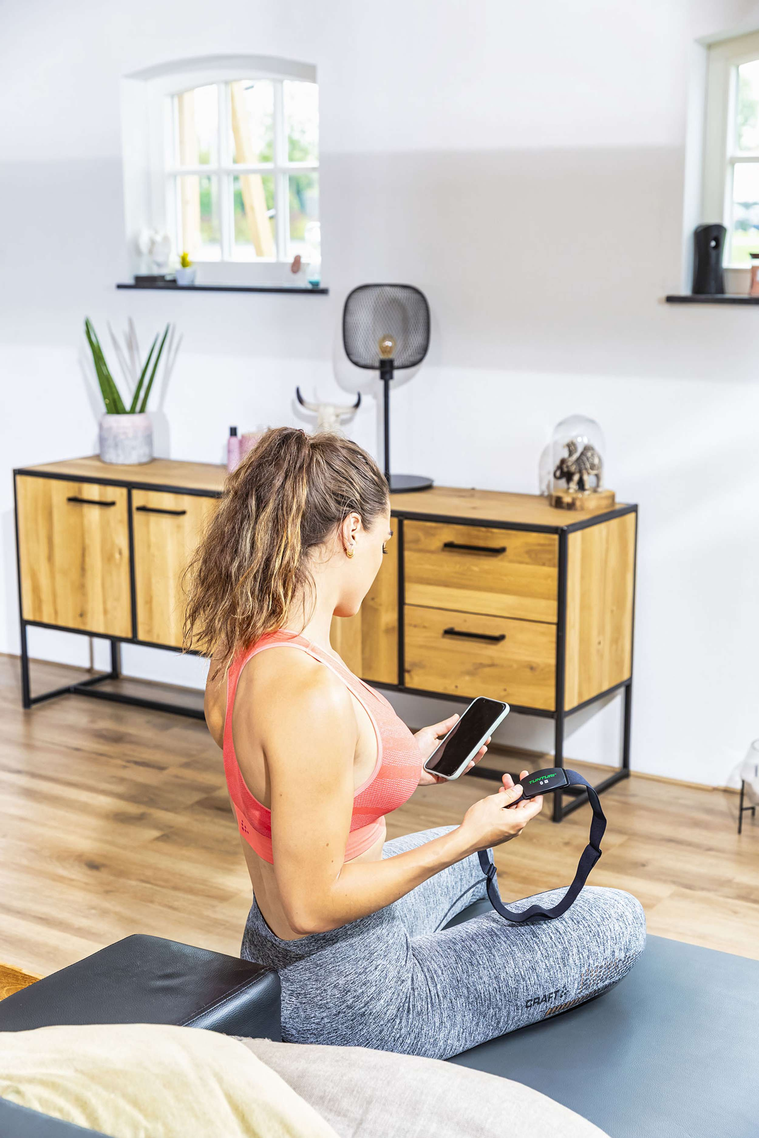 Woman in sportswear sits, holding phone and heart rate monitor. Wooden cabinet, window, and decor in the background. 