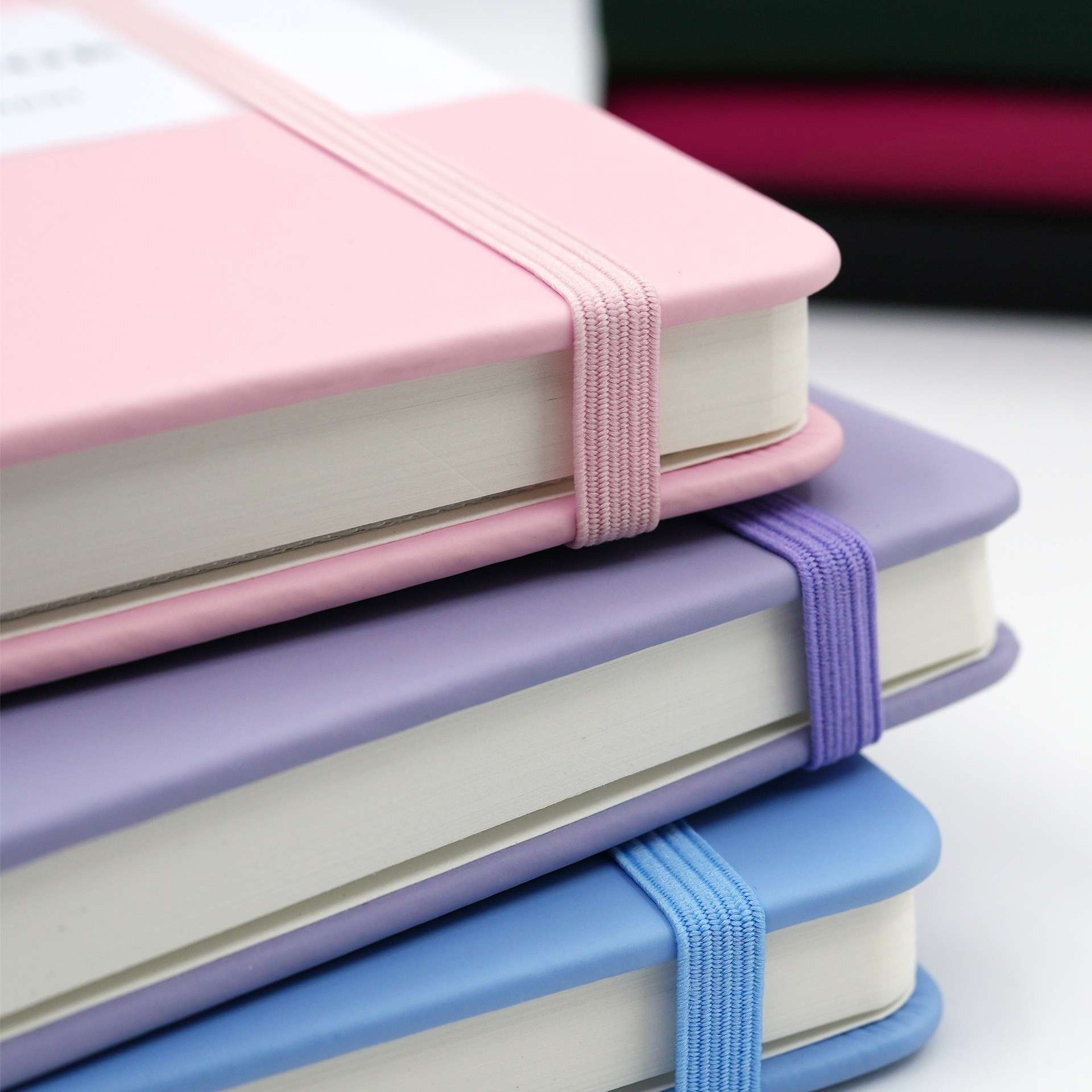 Drei gestapelte Notizbücher mit Gummibändern. Rosa, lila und blaue Notizbücher sind sichtbar. Weiße Seiten und ein weißer Hintergrund.