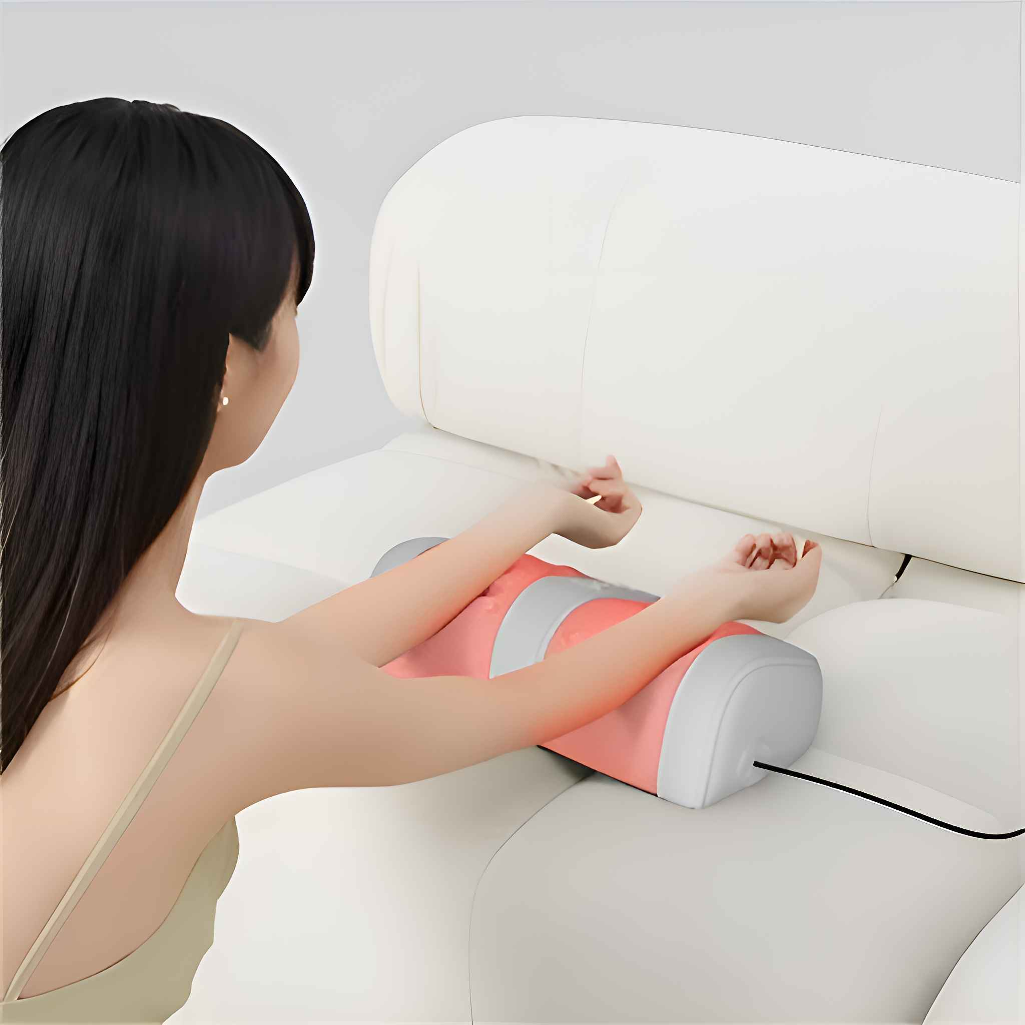 Eine Frau benutzt ein rosa-graues Armmassagegerät auf einem weißen Sofa. Ihre Arme ruhen auf dem Gerät, ein schwarzes Kabel ist angeschlossen.