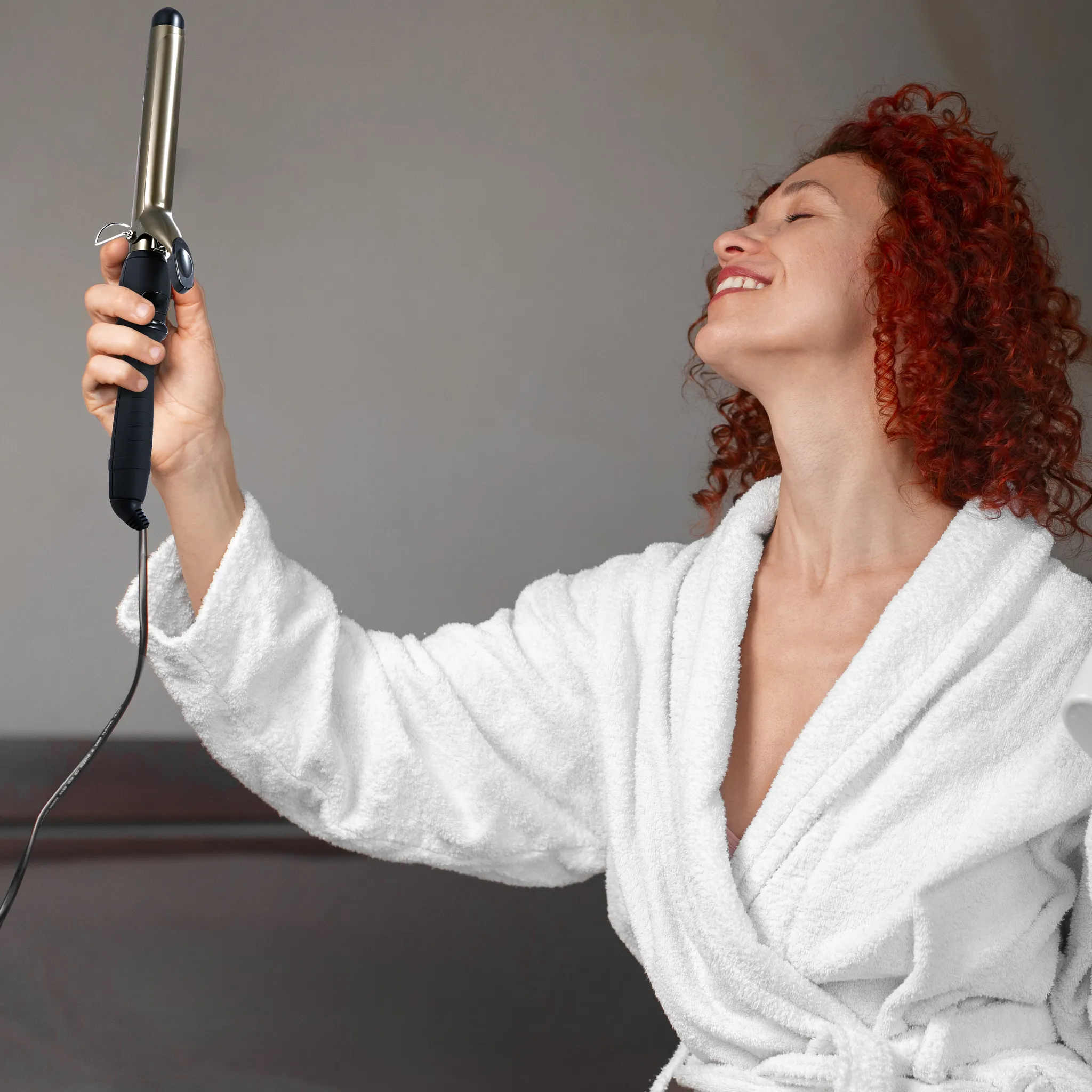 Frau mit roten Locken in einem weißen Bademantel, hält einen Lockenstab. Sie lächelt mit geschlossenen Augen.