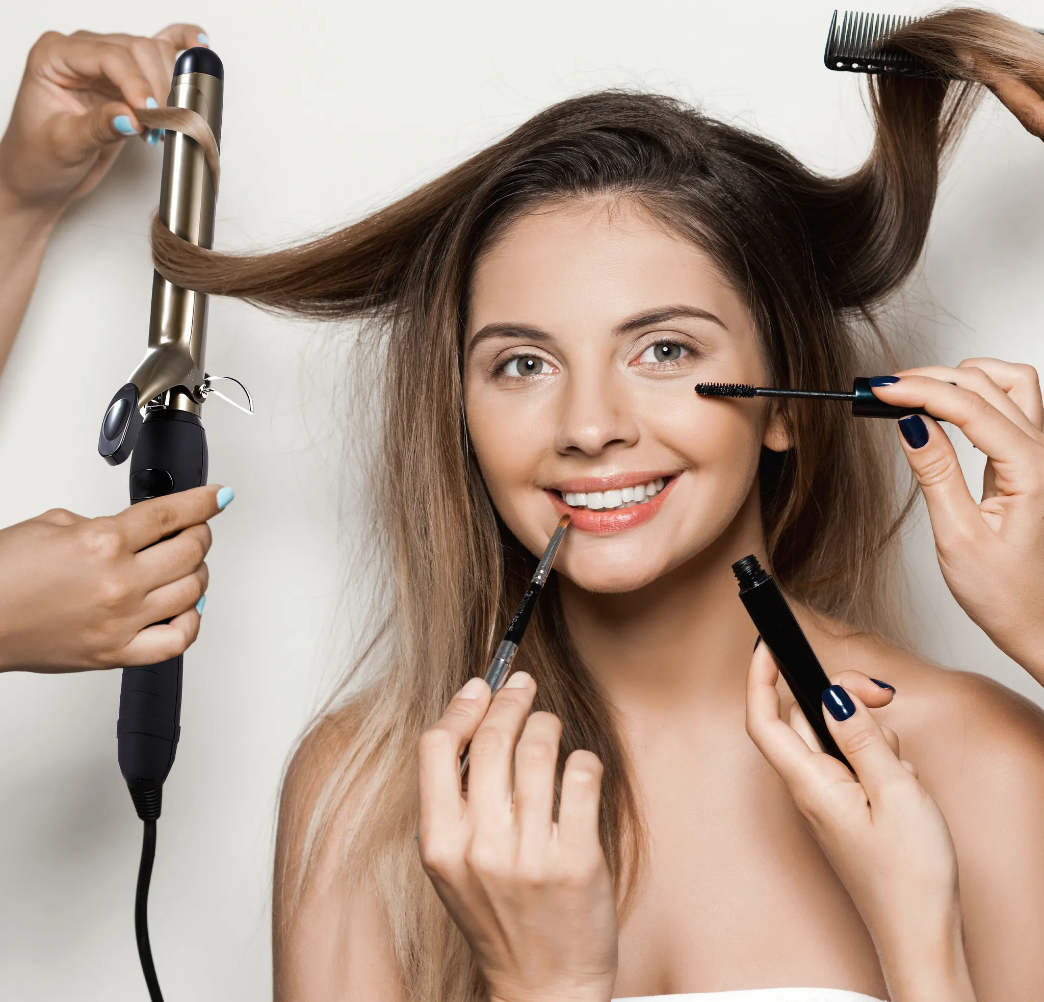 Frau lächelt, während sie geschminkt wird. Hände halten Mascara, Lippenstift und Lockenstab. Haare werden gekämmt.