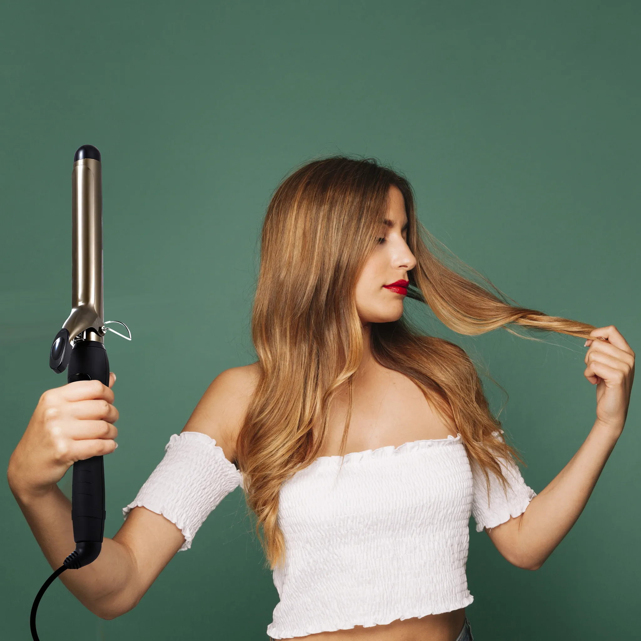 Eine Frau mit langen Haaren hält einen Lockenstab. Sie trägt ein weißes Oberteil und berührt ihr Haar. Grüner Hintergrund.
