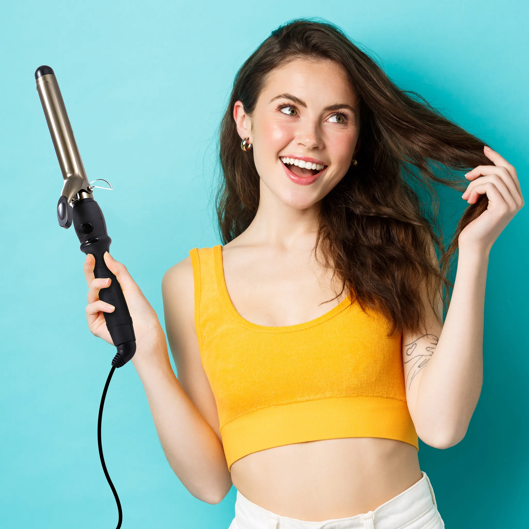 Lächelnde Frau mit Lockenstab. Sie hält Haare, trägt ein gelbes Oberteil, vor blauem Hintergrund.