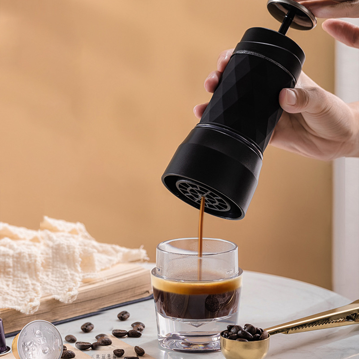 Eine Person gießt Kaffee aus einem schwarzen tragbaren Espressomaschine in ein klares Glas. Kaffeebohnen und ein Löffel liegen auf dem Tisch.