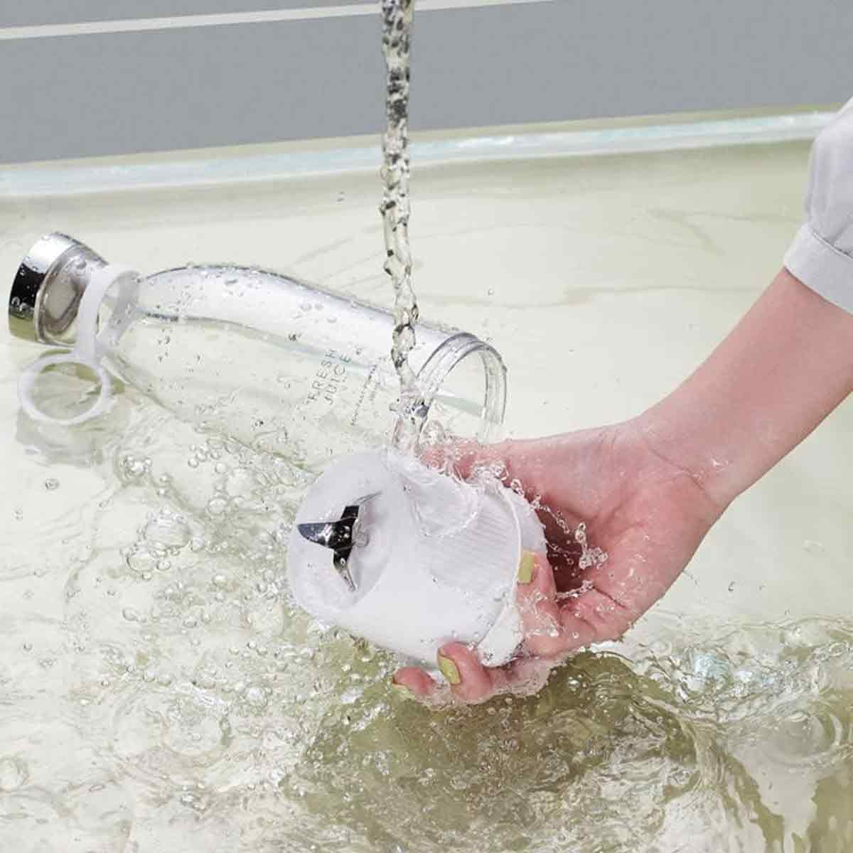 Eine Hand hält einen weißen Mixerfuß unter fließendem Wasser, daneben eine klare Flasche. Wasser spritzt herum und erzeugt eine nasse Szene.