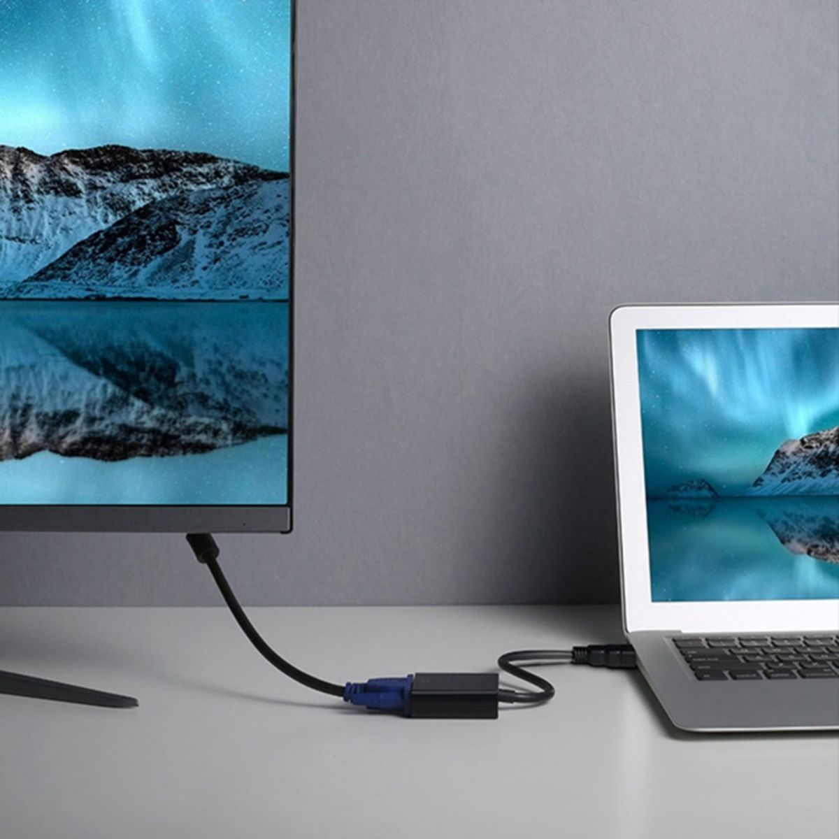 Ein Laptop, der über einen schwarzen Adapter mit einem Monitor verbunden ist. Beide Bildschirme zeigen eine Landschaft mit Bergen.