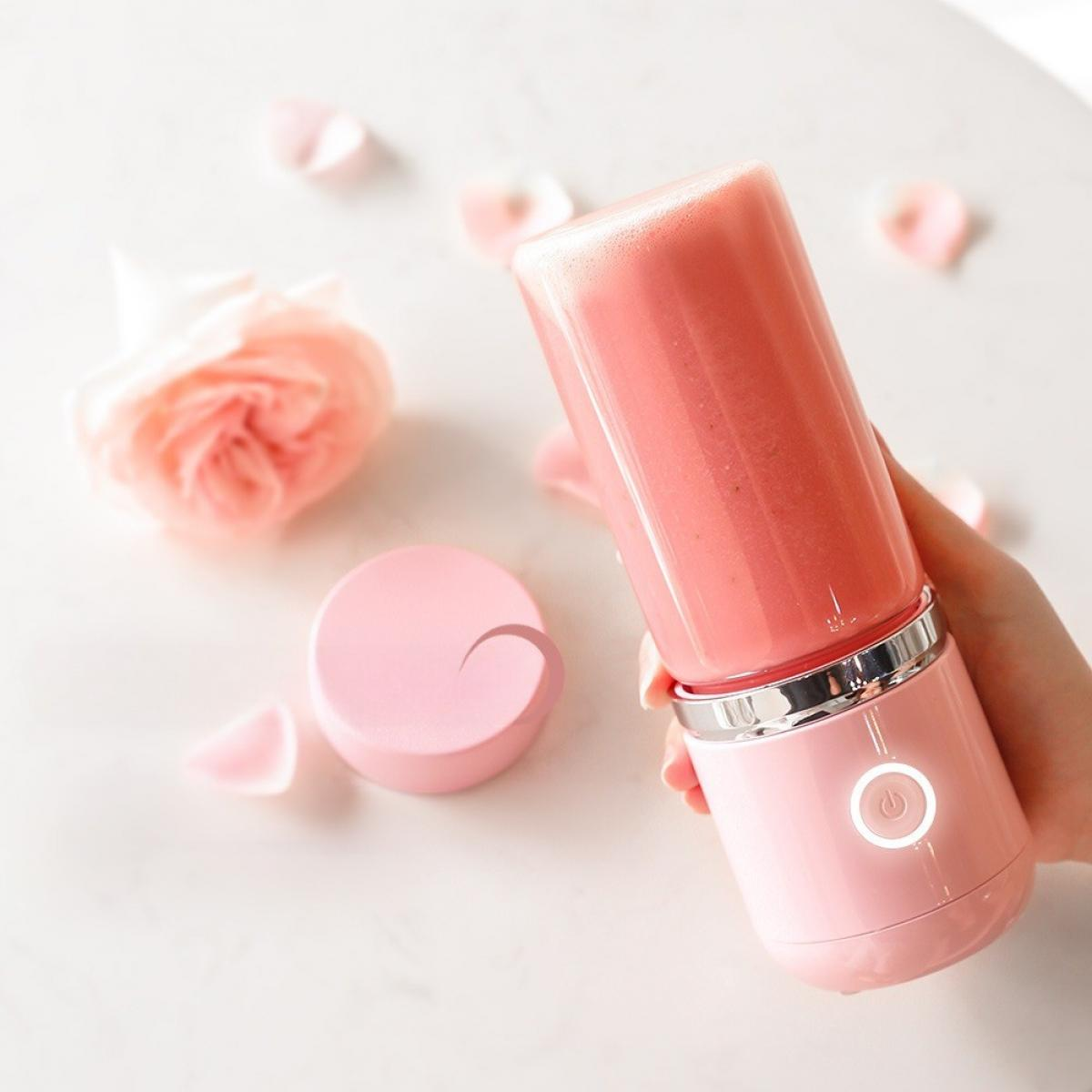 Eine Hand hält einen rosa Mixer mit rosa Smoothie. Rosa Rose, Blütenblätter und ein rosa Deckel liegen auf der weißen Oberfläche.
