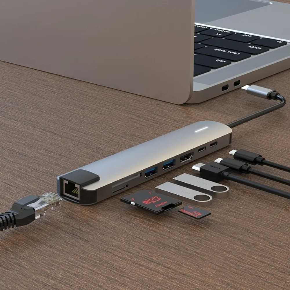 Ein silberner USB-Hub, der mit einem Laptop verbunden ist. Verschiedene Anschlüsse und angeschlossene Kabel sind sichtbar, zusammen mit SD-Karten und USB-Laufwerken.