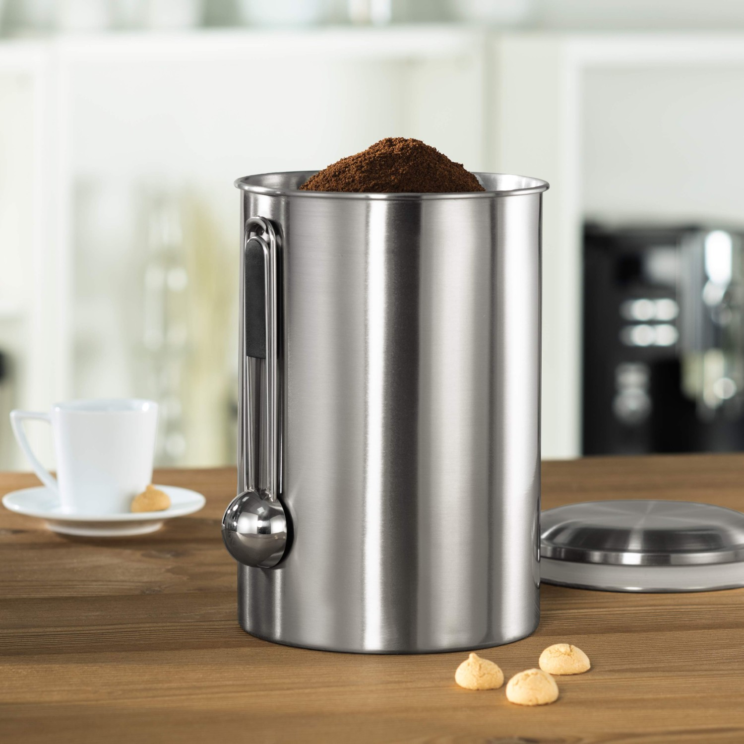 Edelstahlbehälter mit Kaffeepulver, Löffel befestigt. Daneben eine Tasse, Untertasse, Kekse auf einem Holztisch.