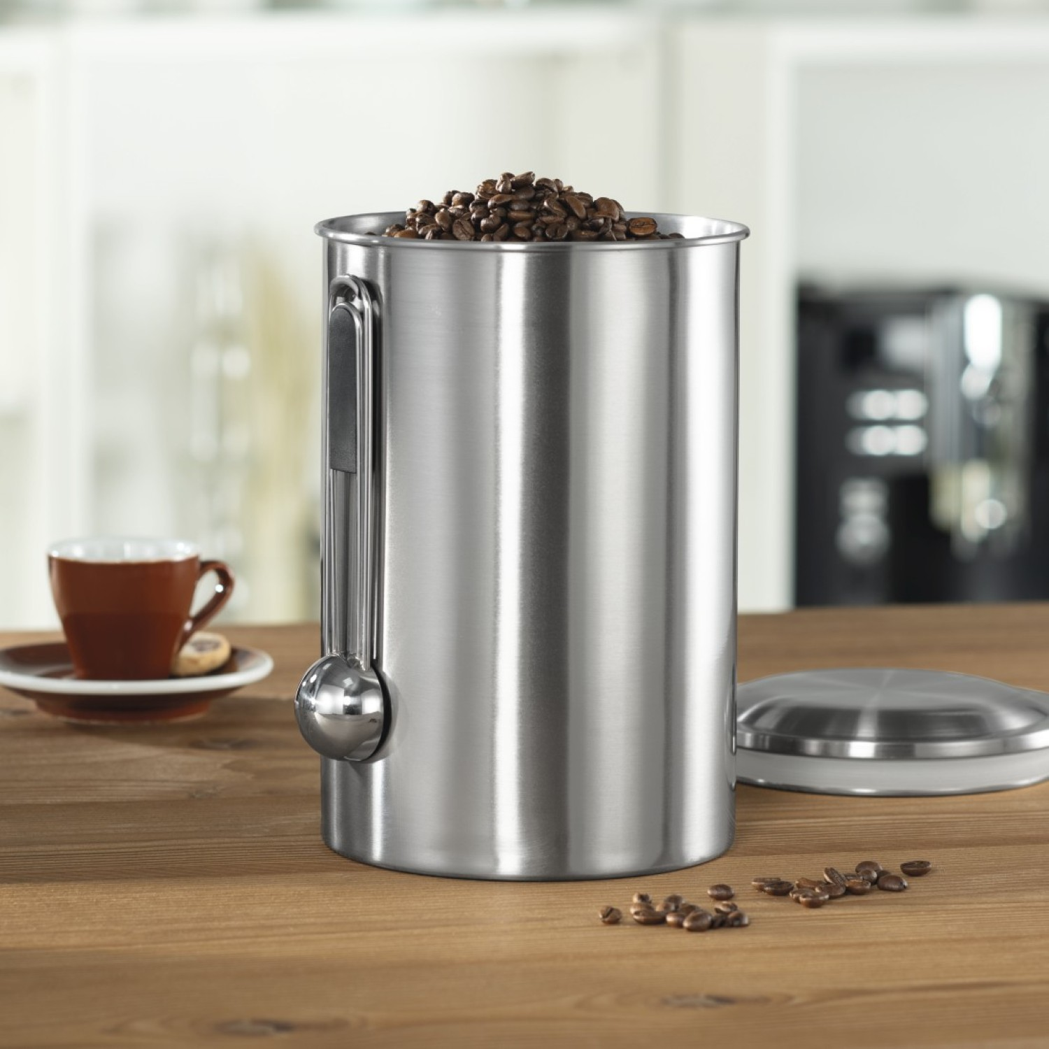 Edelstahl-Kaffeebehälter gefüllt mit Kaffeebohnen. Links sind eine Tasse und Untertasse. Bohnen sind auf dem Holztisch verstreut.