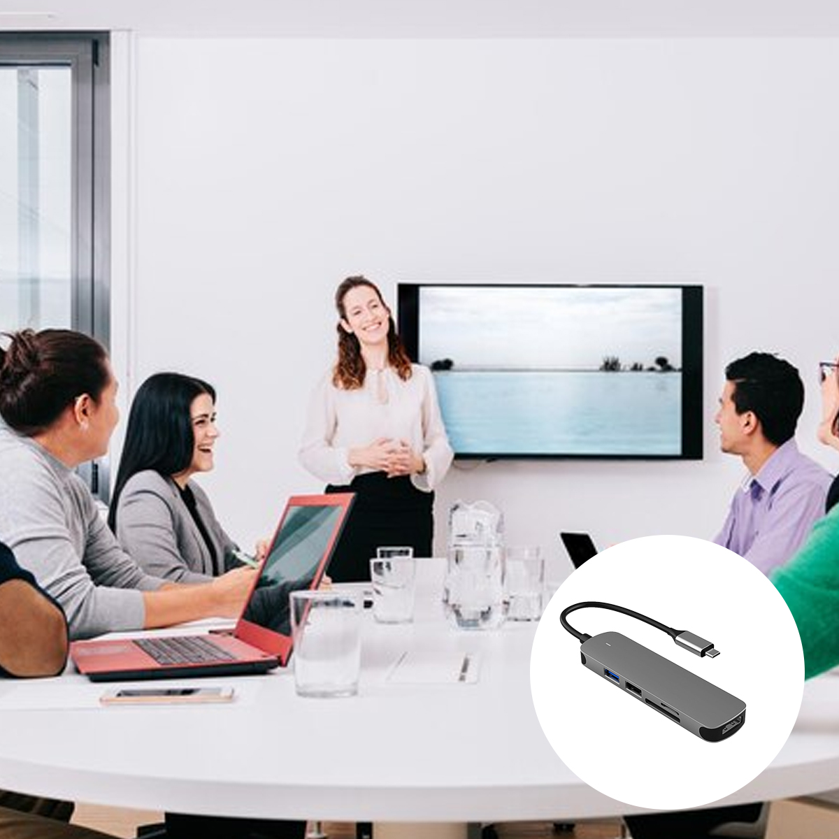 Ein Geschäftstreffen. Eine Frau präsentiert einer Gruppe. Ein Laptop und ein grauer Hub liegen auf dem Tisch.