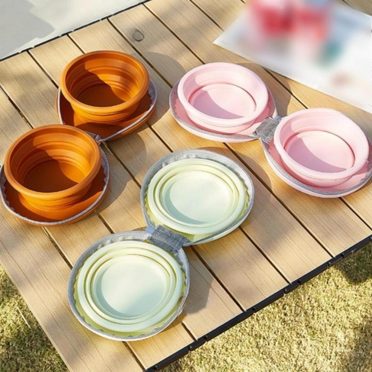 Zusammenklappbare Schüsseln in Orange, Rosa und Grün auf einem Holztisch. Schüsseln sind offen und geschlossen. Der Hintergrund ist Gras.