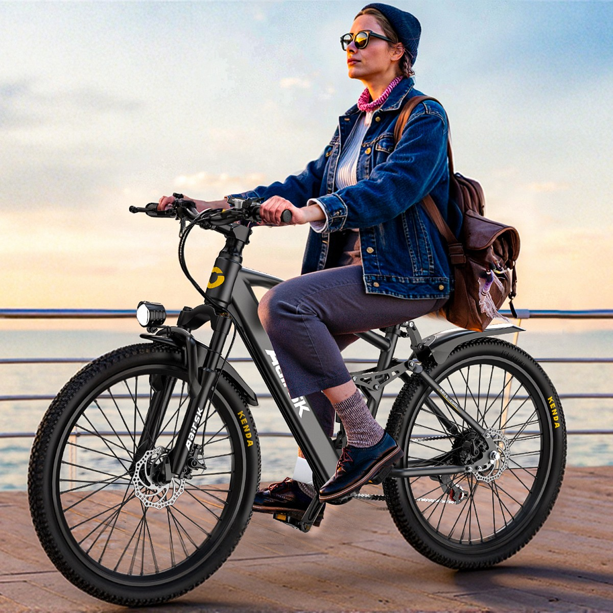 Eine Frau fährt auf einem schwarzen Elektrofahrrad auf einem Pier. Sie trägt eine Jeansjacke, Sonnenbrille und einen Rucksack, mit dem Meer im Hintergrund.