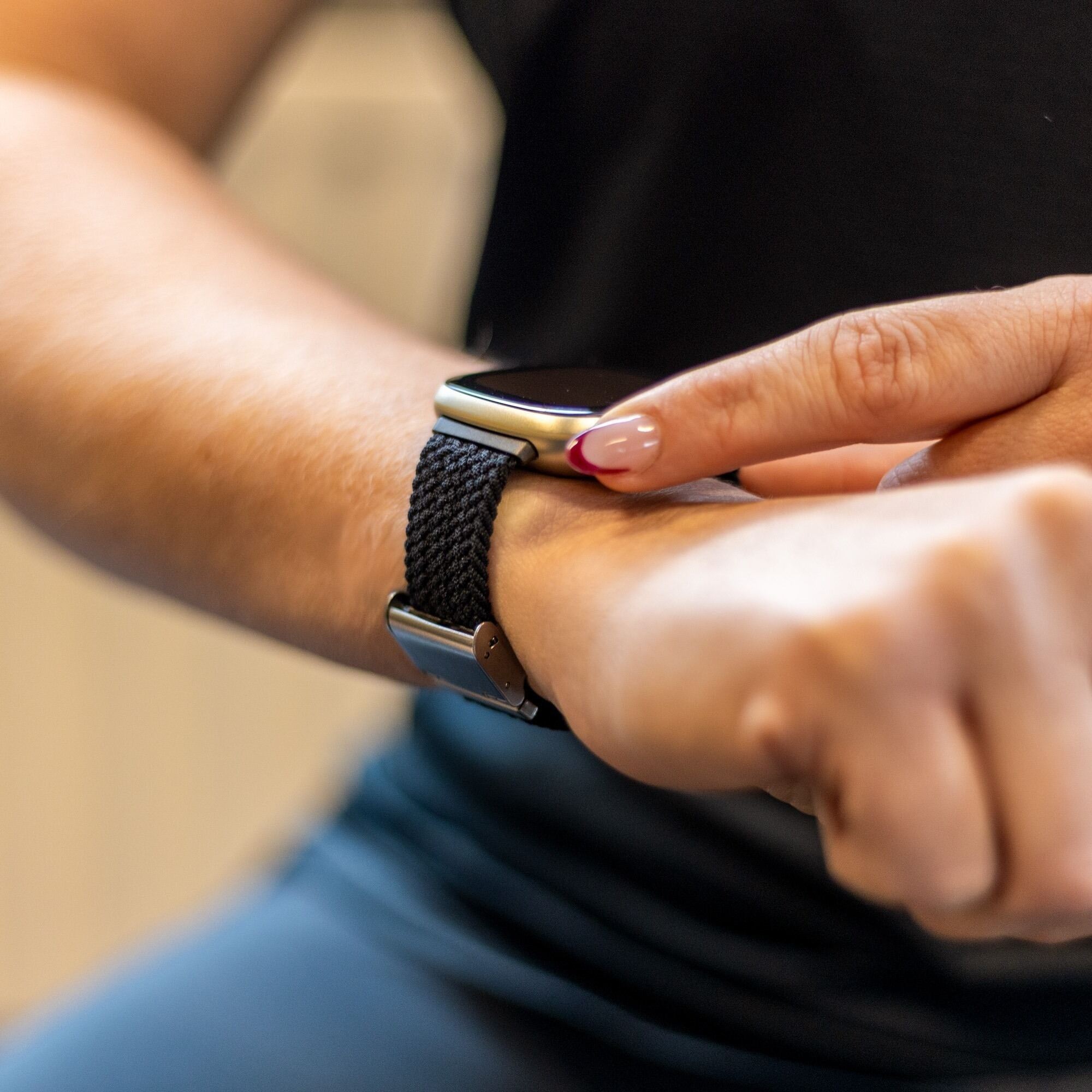 Eine Hand berührt eine Smartwatch. Die Uhr hat ein schwarzes Armband und ein goldenes Gehäuse. Der Nagel hat eine rote Spitze.