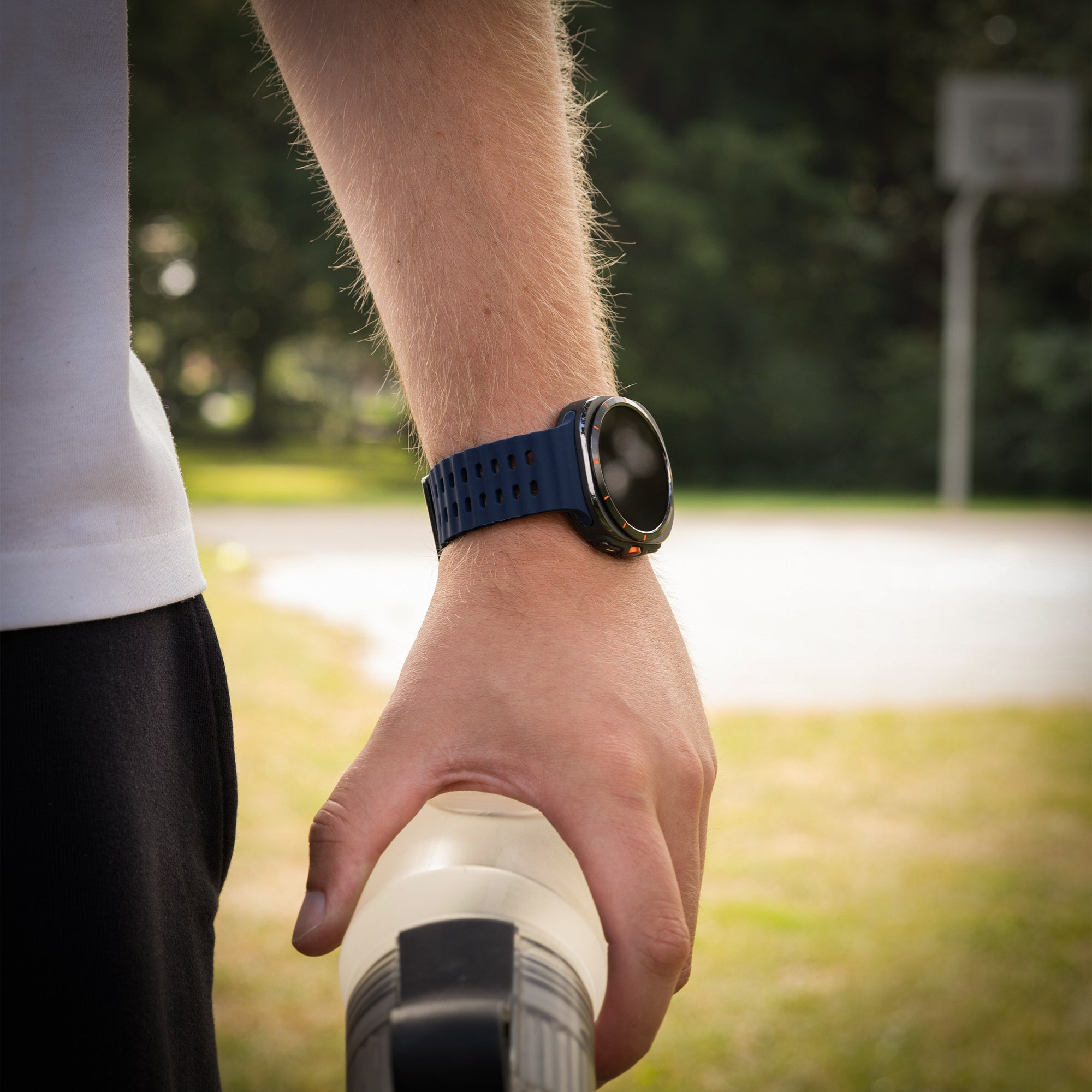 Der Arm einer Person mit einer blauen Smartwatch, die eine Wasserflasche hält. Der Hintergrund zeigt einen Park mit Bäumen und einem Basketballkorb.