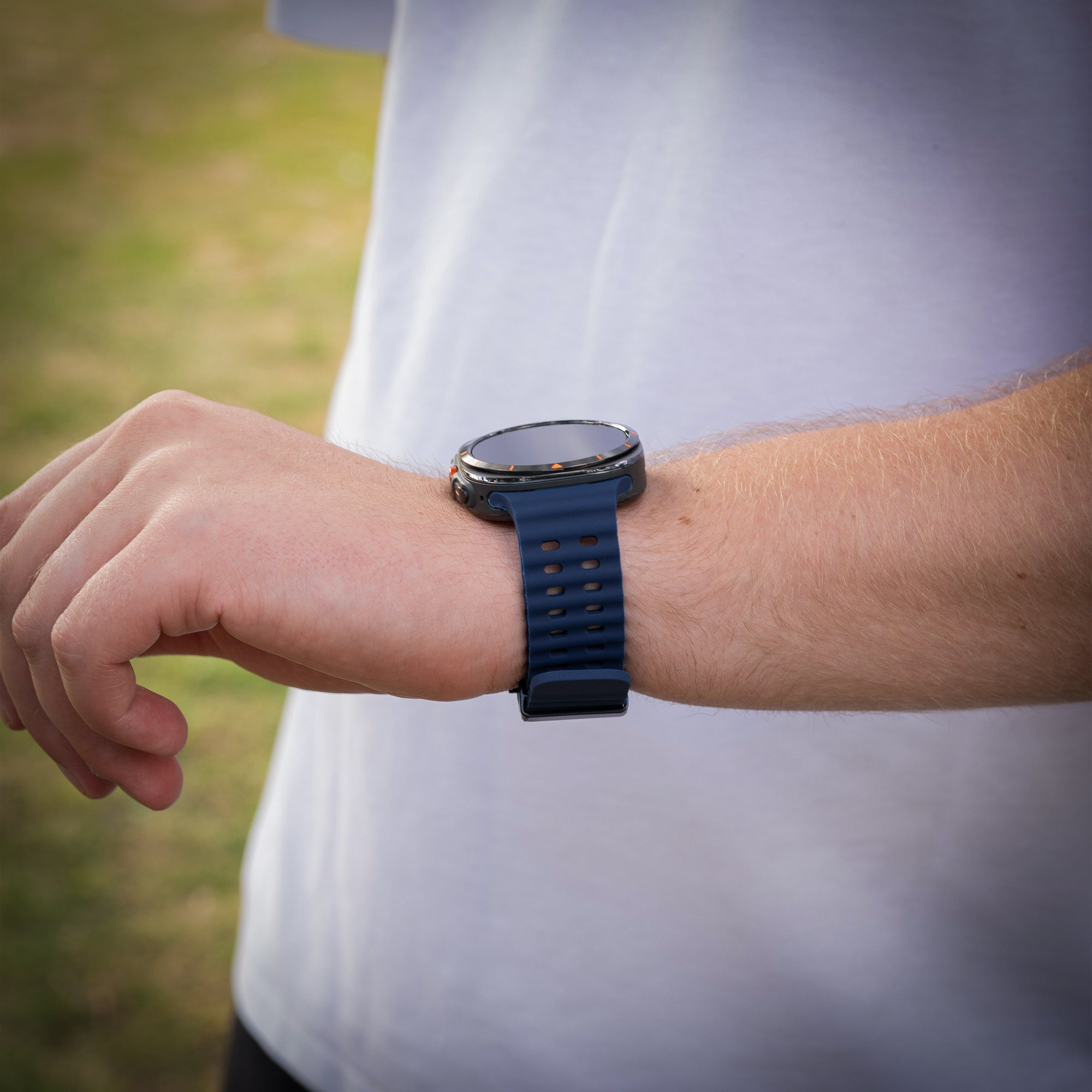 Eine Person trägt eine blaue Smartwatch. Die Person trägt ein weißes T-Shirt. Grünes Gras im Hintergrund.