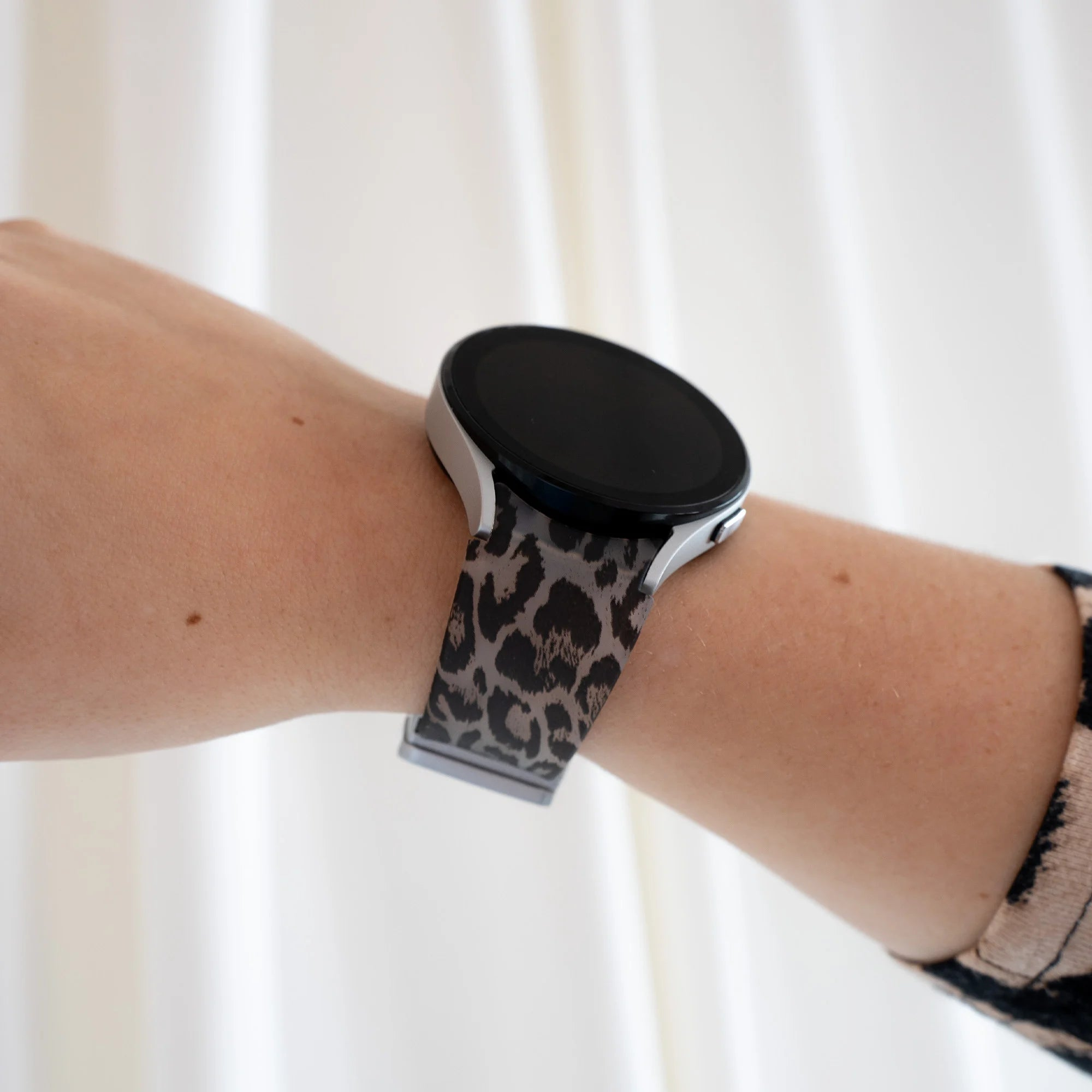 Handgelenk mit silberner Smartwatch. Leopardmuster-Armband. Hintergrund: weißer Vorhang.