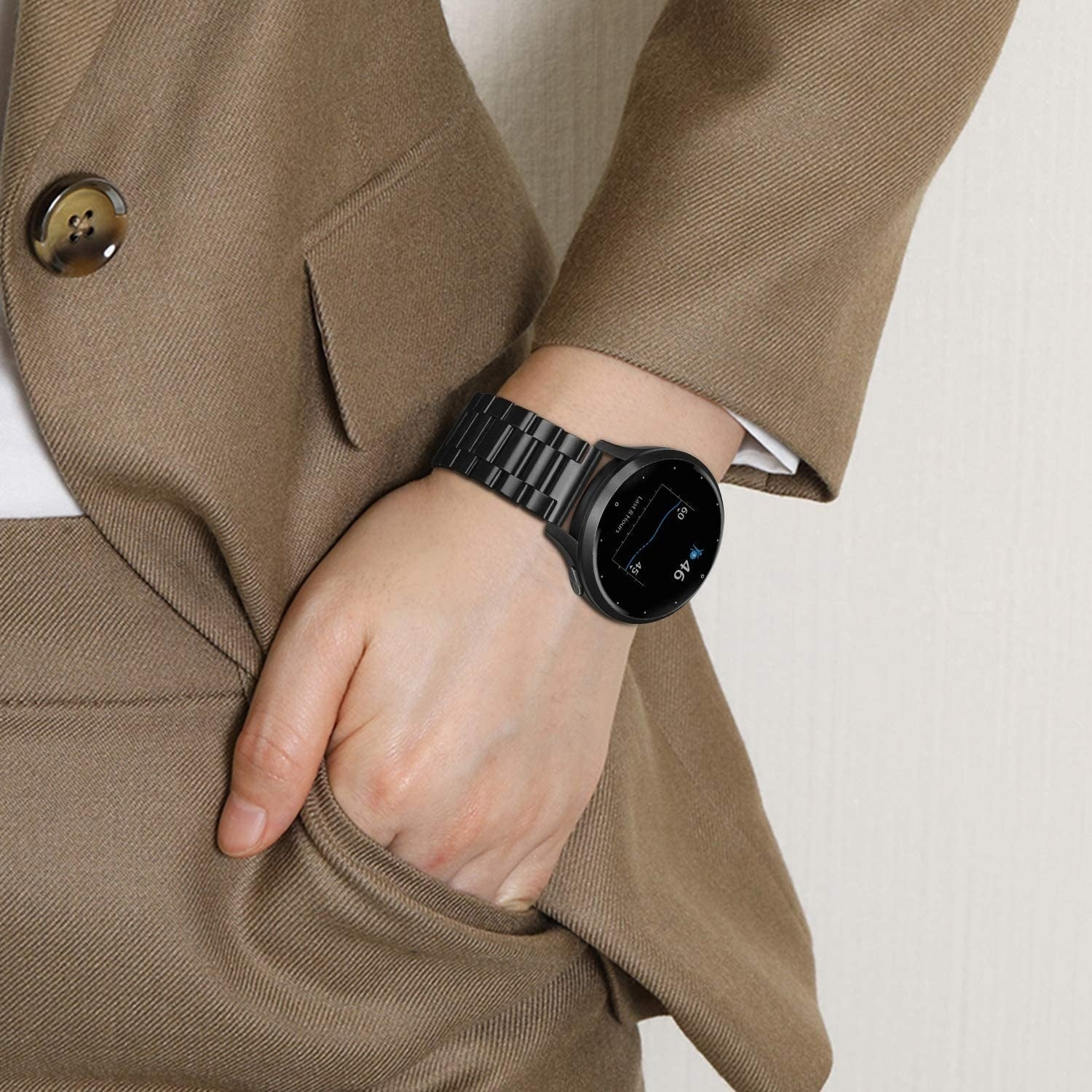Eine Person in einem beigefarbenen Anzug trägt eine schwarze Smartwatch. Die Hand ist in der Tasche. Der Hintergrund ist eine weiße Wand.