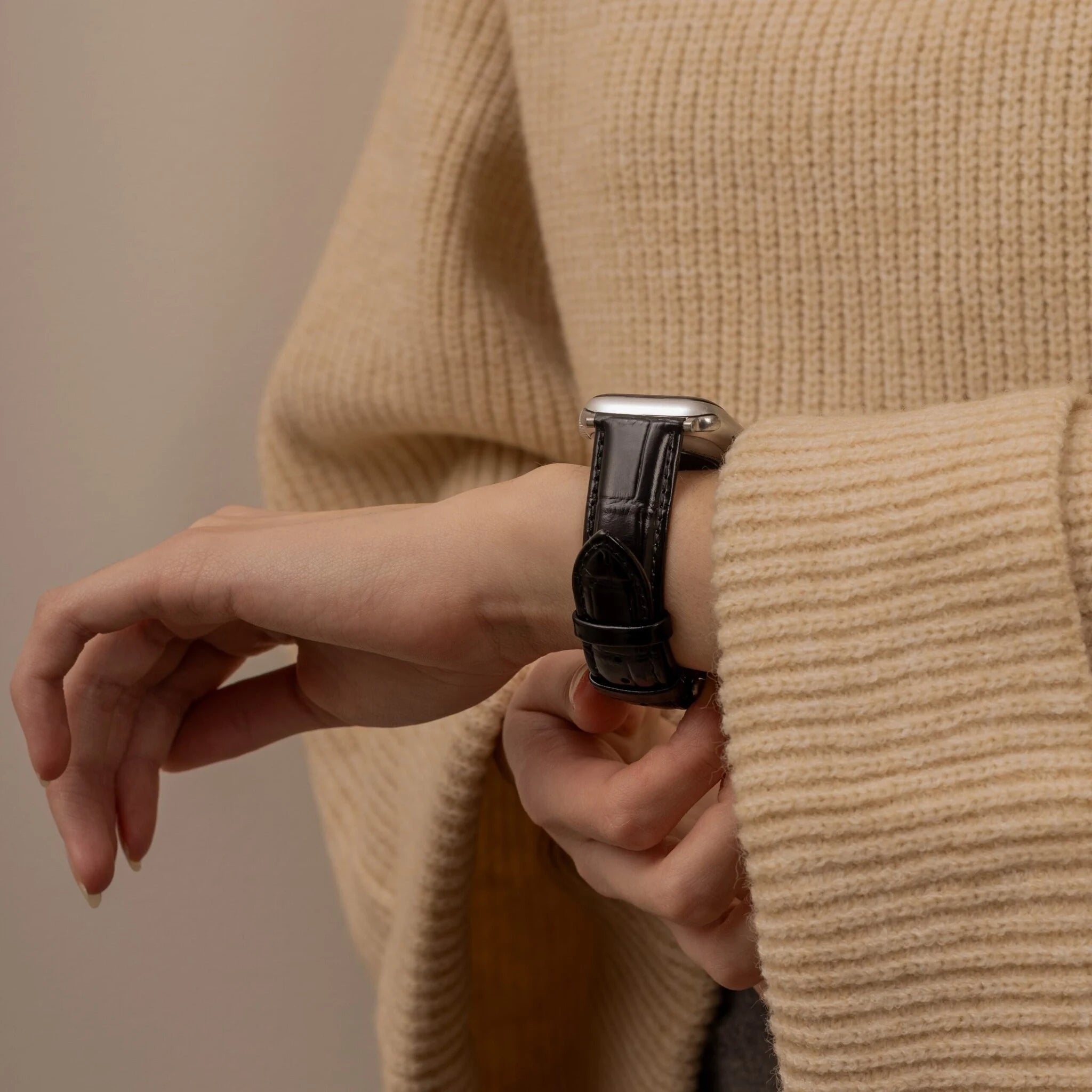 Eine Person trägt einen cremefarbenen Pullover und eine schwarze Lederuhr, das Handgelenk im Fokus. Hintergrund neutral.