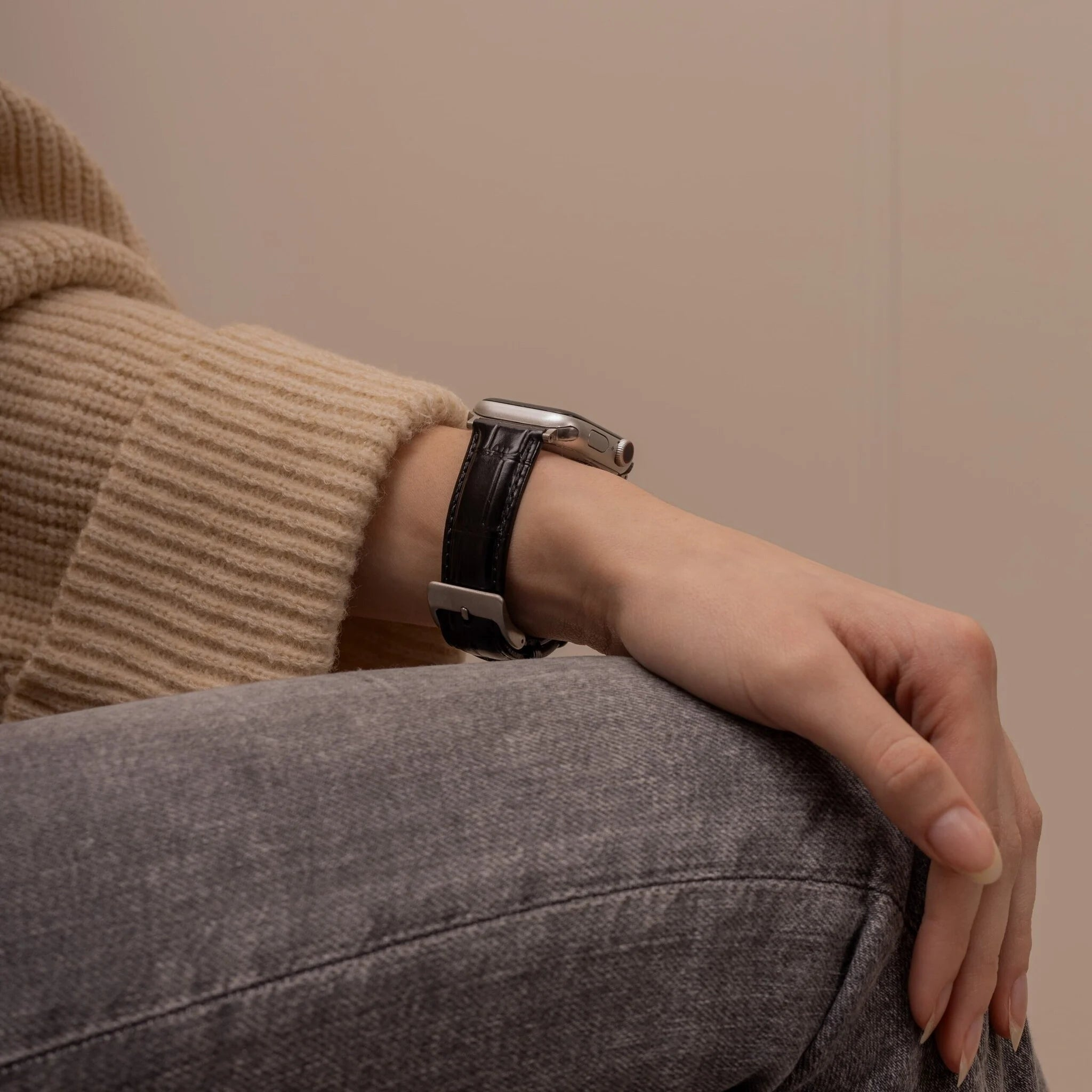 Person trägt eine silberne Uhr mit schwarzem Lederarmband. Beiger Pullover und graue Jeans sind ebenfalls sichtbar.