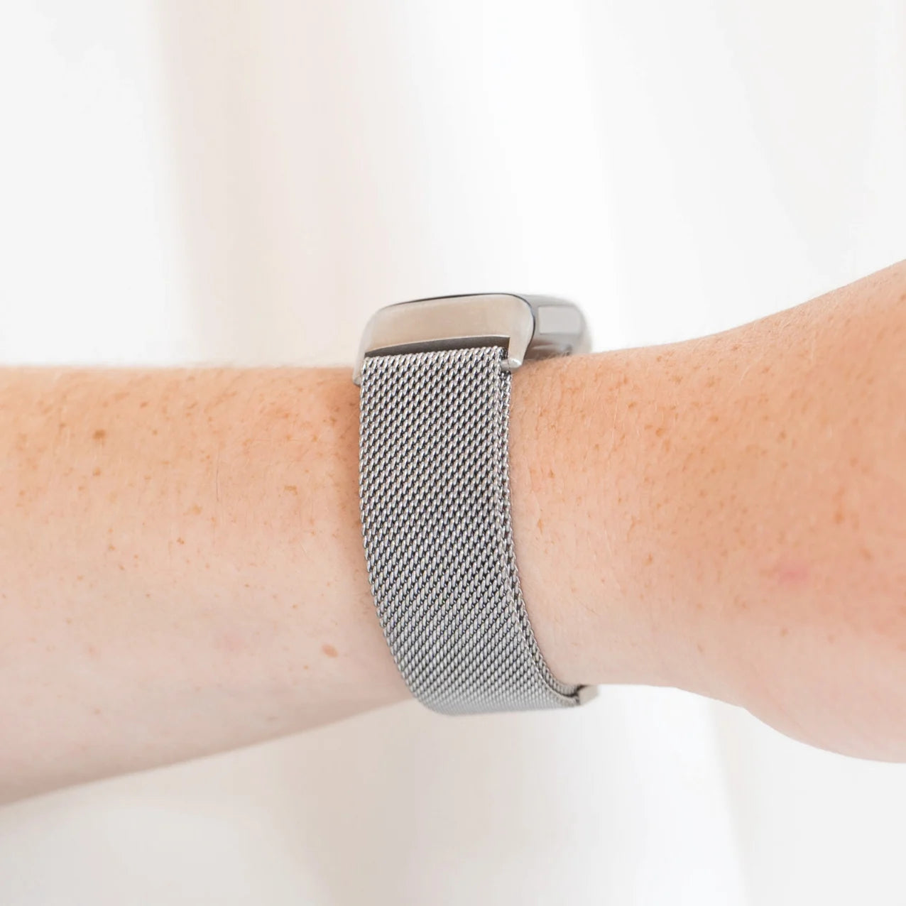 Ein Arm mit einer silbernen Uhr und einem Mesh-Armband. Die Haut ist hell mit Sommersprossen. Der Hintergrund ist ein schlichtes Weiß.