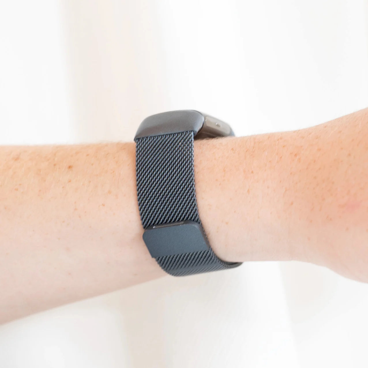 Der Arm einer Person mit einer grauen Smartwatch. Die Uhr hat ein Mesh-Armband. Die Haut ist hell mit Sommersprossen.