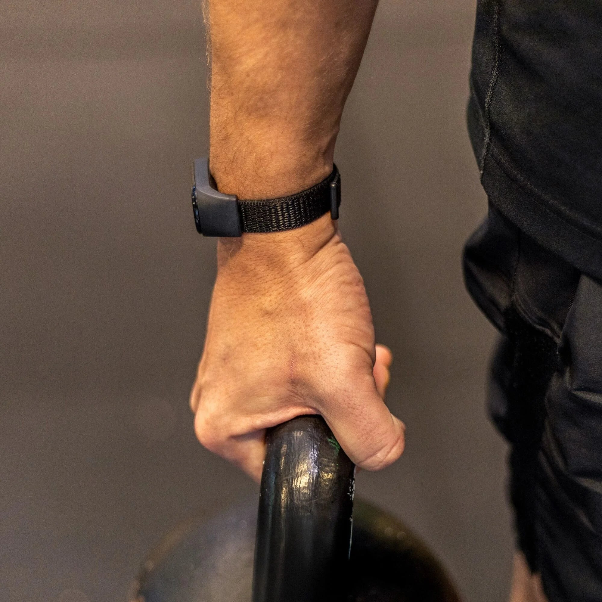 Eine Männerhand hält eine schwarze Kettlebell. Er trägt eine Uhr und ein schwarzes Hemd. Der Hintergrund ist dunkelgrau.