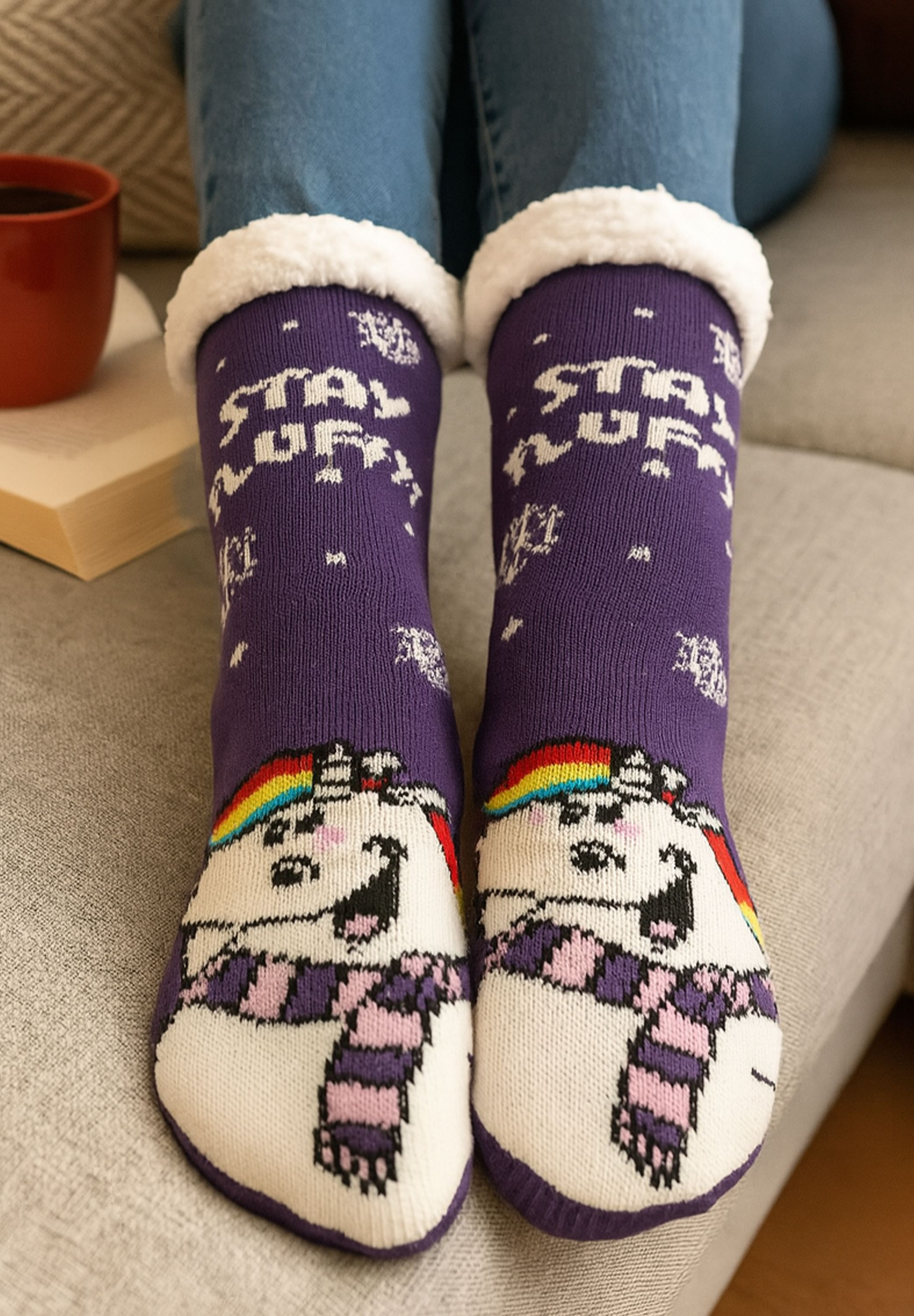 Person trägt lila Socken mit Einhorn-Design, weißem, flauschigem Oberteil und der Aufschrift 'STAY UP'. Im Hintergrund sind eine rote Tasse und ein Buch zu sehen.