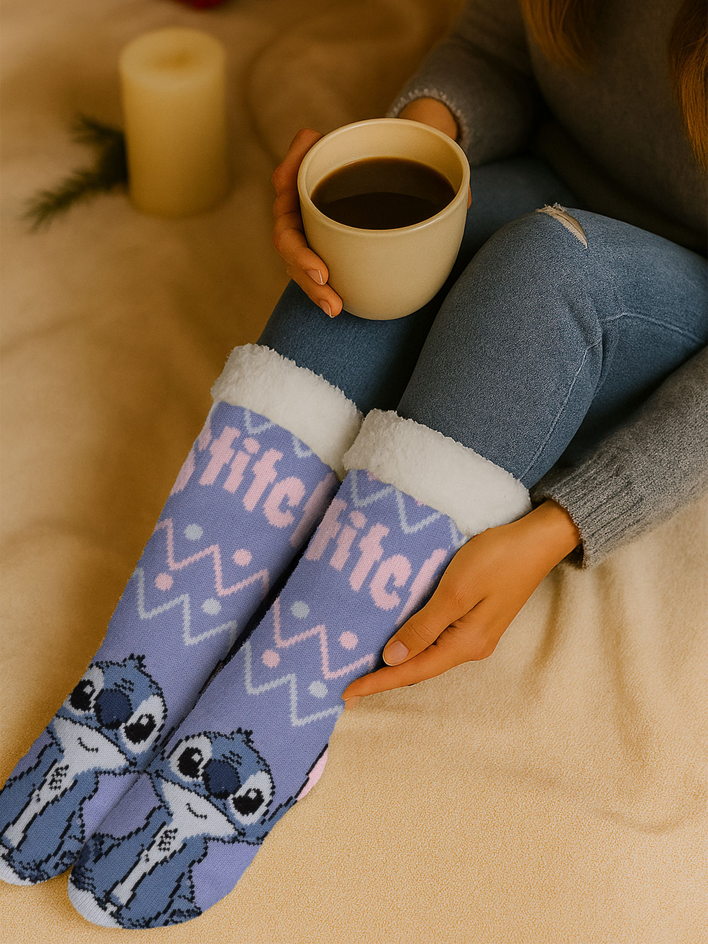 Person trägt Stitch-Socken und hält Kaffee. Hintergrund: Kerze, Decke.