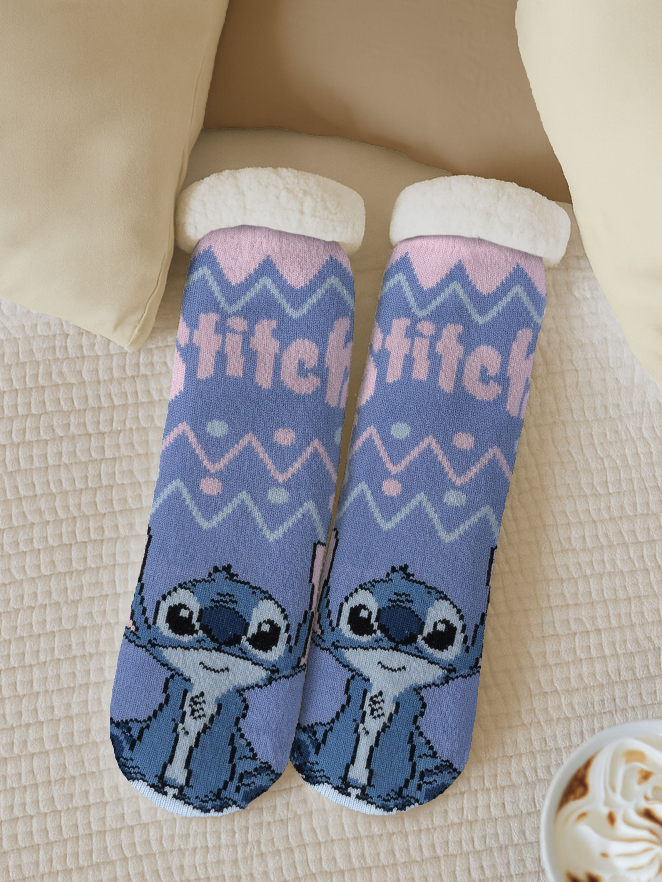 Zwei Stitch-Socken liegen auf einem Bett. Sie sind blau mit rosa Akzenten. Im Hintergrund sind Kissen und eine Kaffeetasse zu sehen.