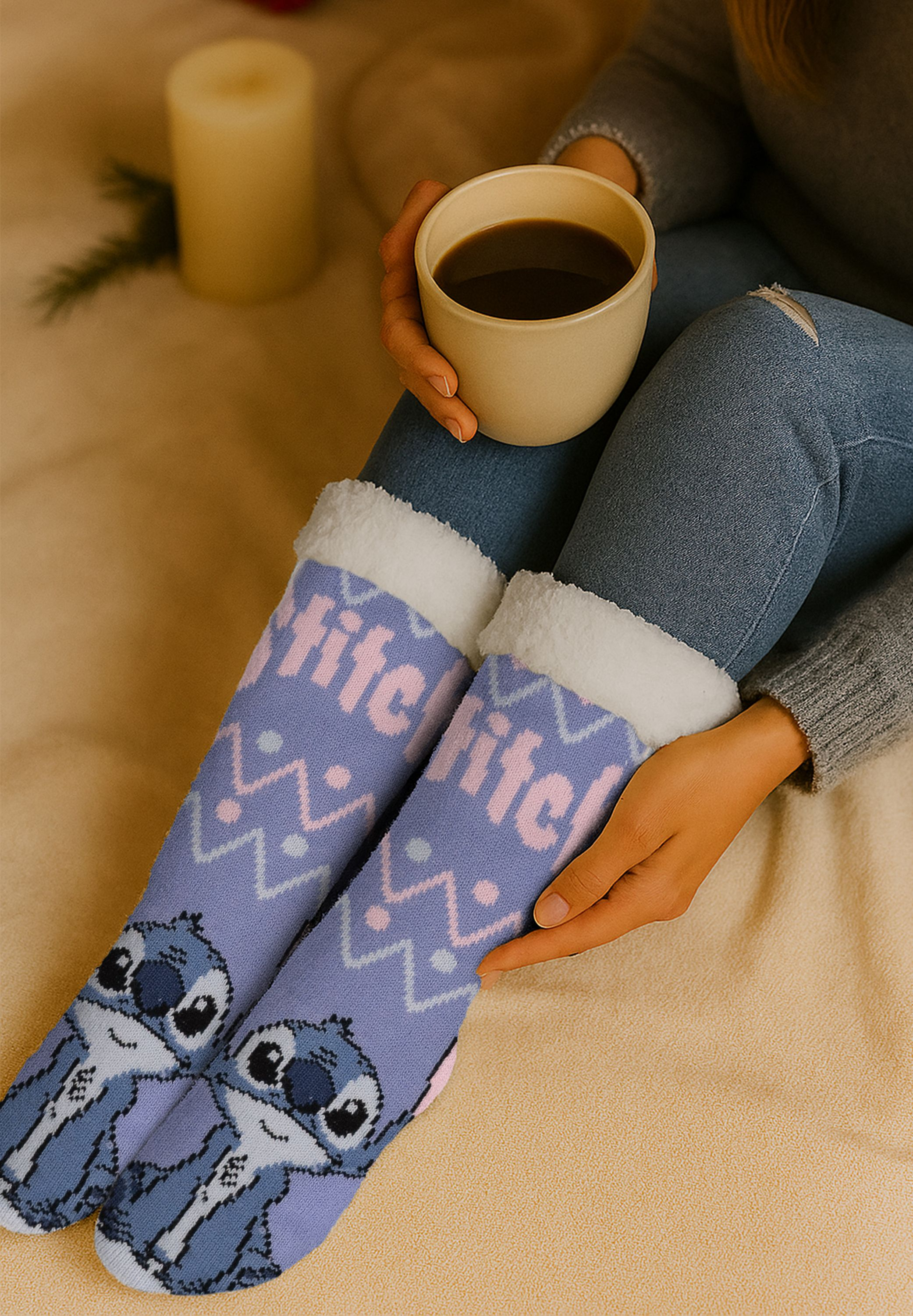 Person hält einen Kaffeebecher, trägt Stitch-Socken, mit einer Kerze daneben. Jeans und ein Pullover sind ebenfalls sichtbar.